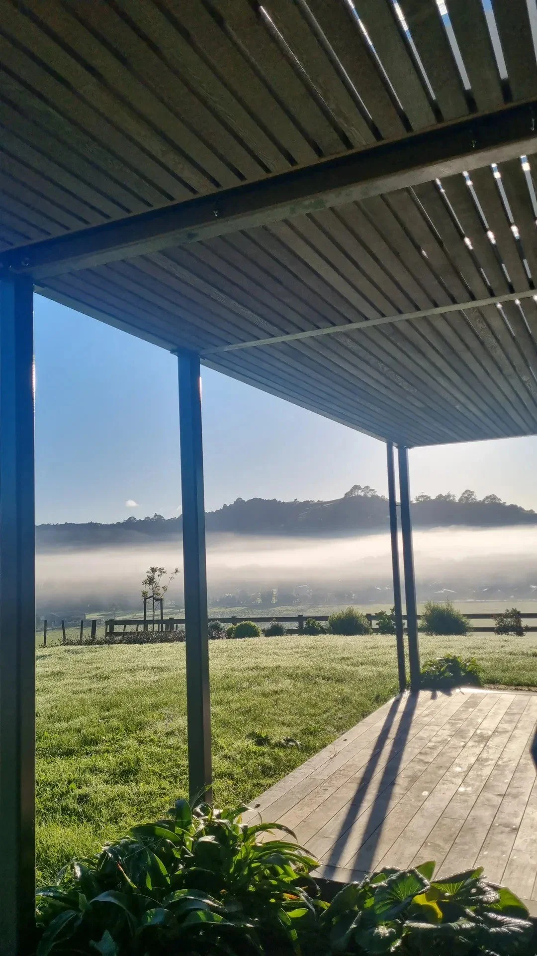 View from under a covered outdoor patio looking out over a grassy yard, misty hills, and cloudy sky in the distance.