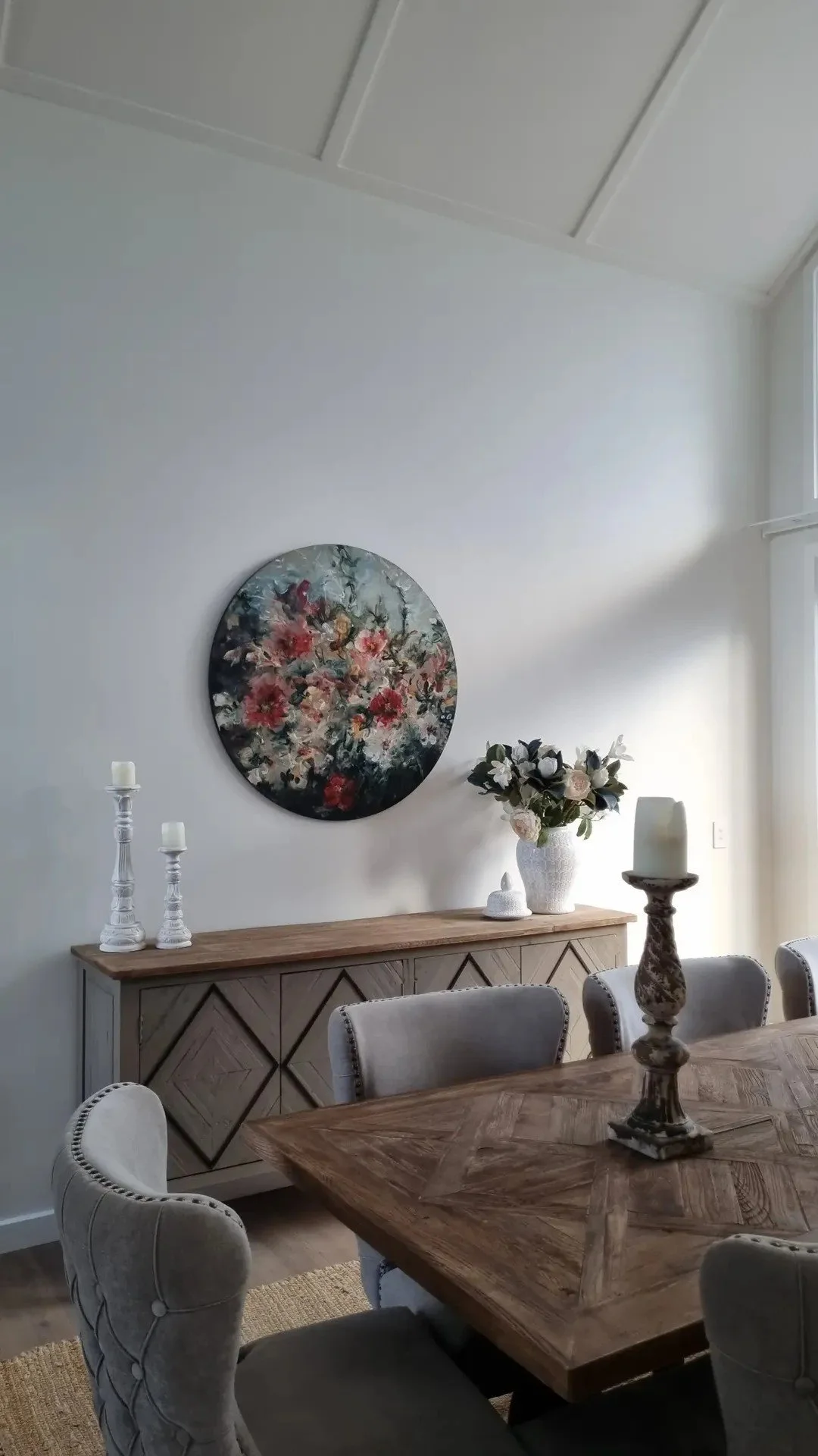 A dining room with a wooden table, beige upholstered chairs, a wooden sideboard with decorative candles, a floral painting on the wall, a vase with flowers, and candleholders.