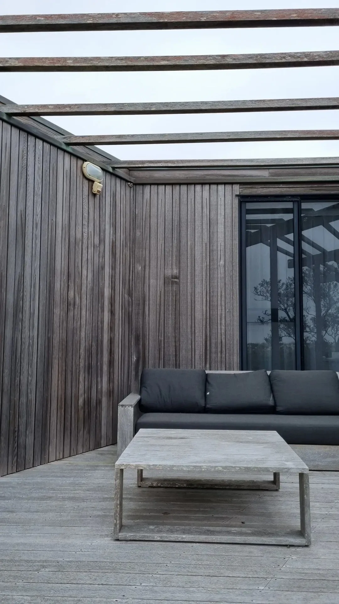Outdoor patio area with dark wooden walls, gray cushioned sofa, and concrete coffee table. Overcast sky visible above.
