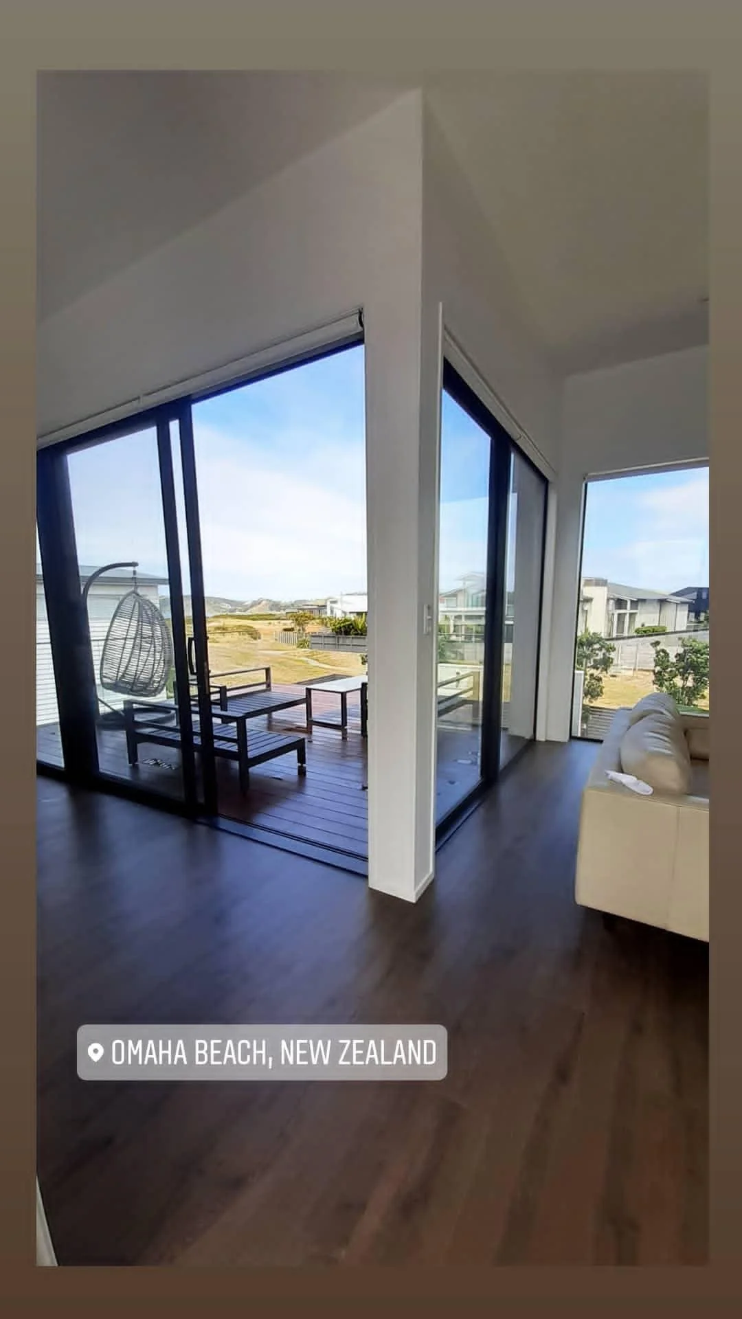 Interior of a modern home with large glass sliding doors leading to an outdoor deck with outdoor furniture, overlooking a beach view in Omaha Beach, New Zealand.
