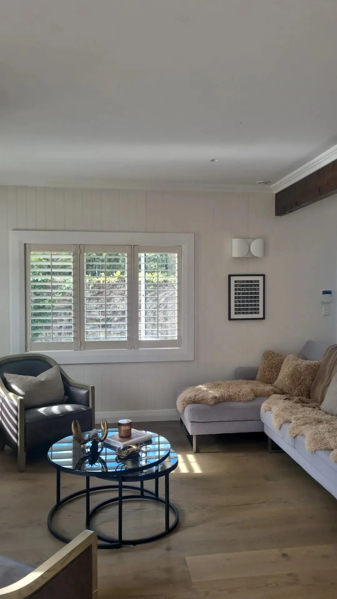 Living room with beige sofa, black armchair, round glass coffee table, window with white shutters, and decorative wall art.