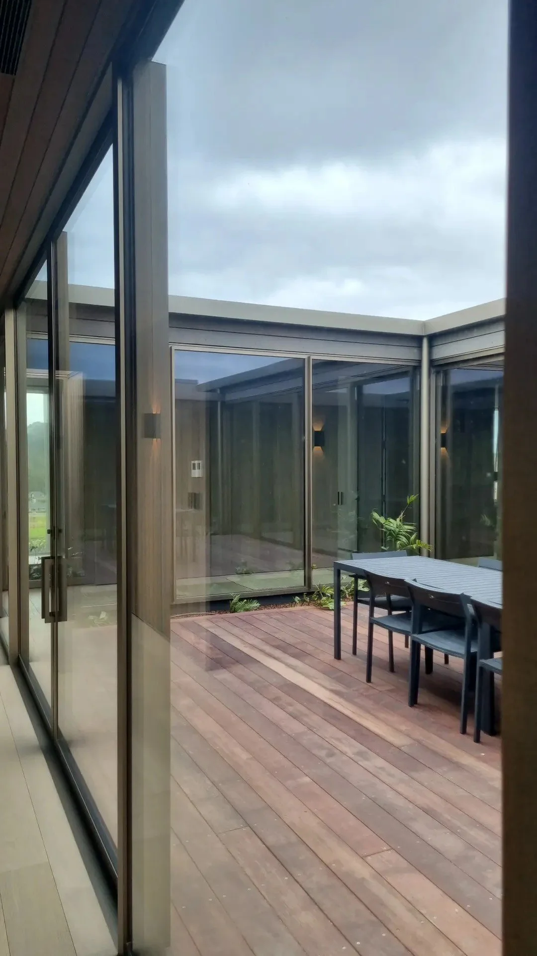 View of a modern outdoor patio with wooden flooring, a rectangular table, chairs, and glass walls, under a cloudy sky.