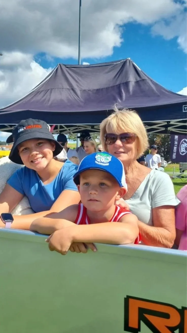 Three people, an older woman and two young children, are smiling and posing in front of a tent at an outdoor event on a partly cloudy day.