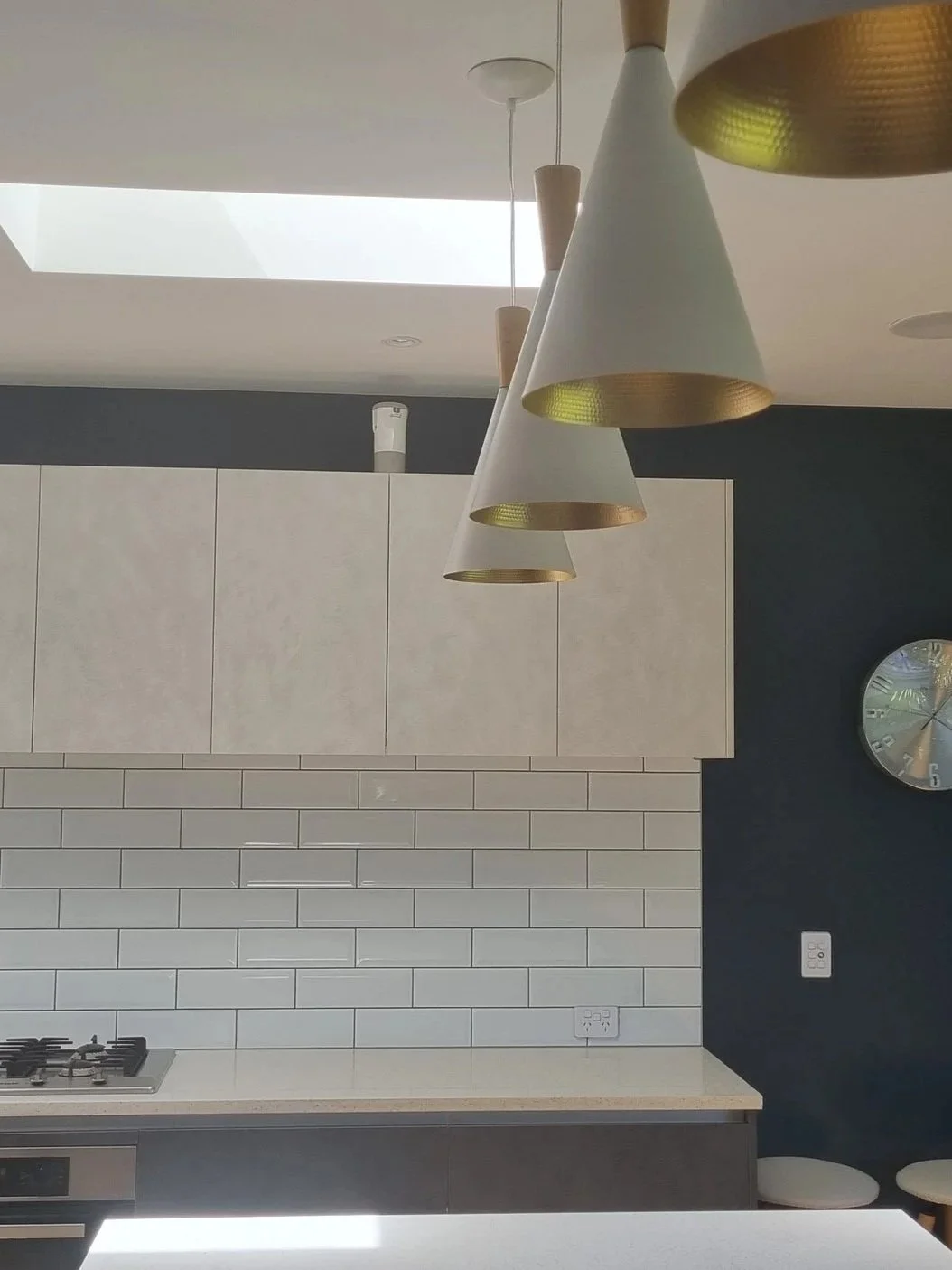 Contemporary kitchen with hanging cone-shaped pendant lights, white cabinetry, a black accent wall, white subway tile backsplash, a built-in stove, a wall clock, and a skylight.