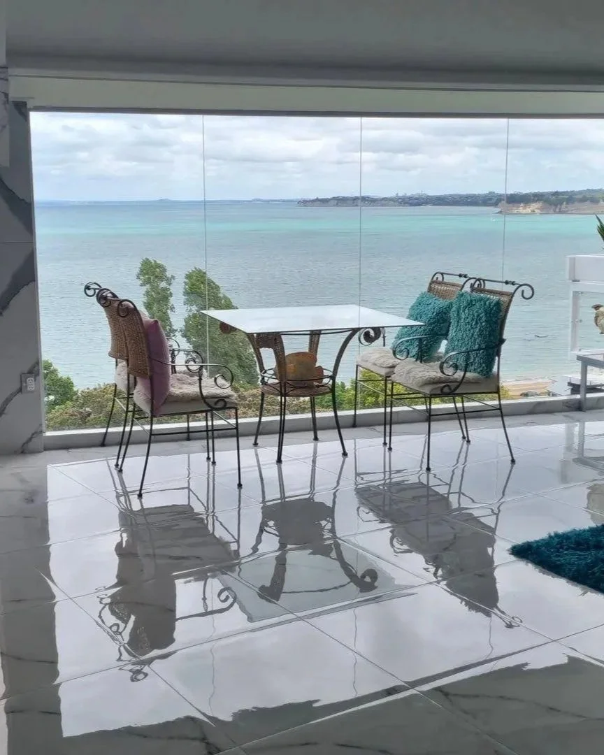Balcony with four ornate metal chairs and a small white table, overlooking a large body of water with a cloudy sky and distant shoreline.