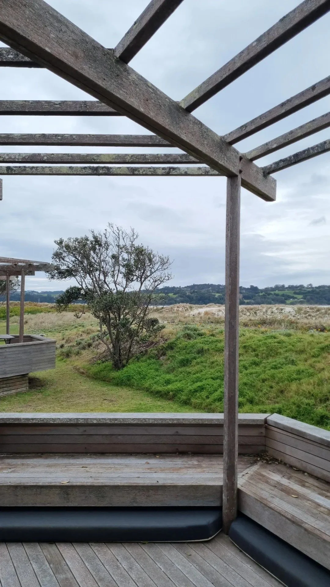 View from a wooden balcony overlooking a grassy landscape with a tree and distant hills under an overcast sky.