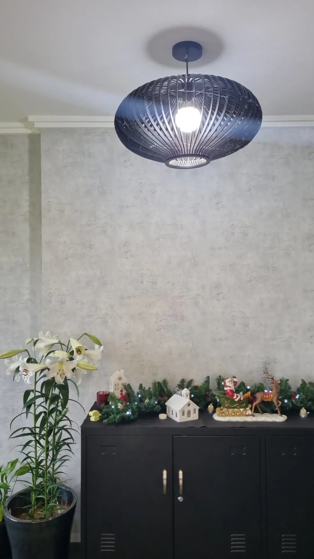 A modern pendant light fixture hangs from the ceiling above a black cabinet decorated with Christmas and holiday items. To the left, a potted plant with white lilies. The wall behind is plain and light-colored.