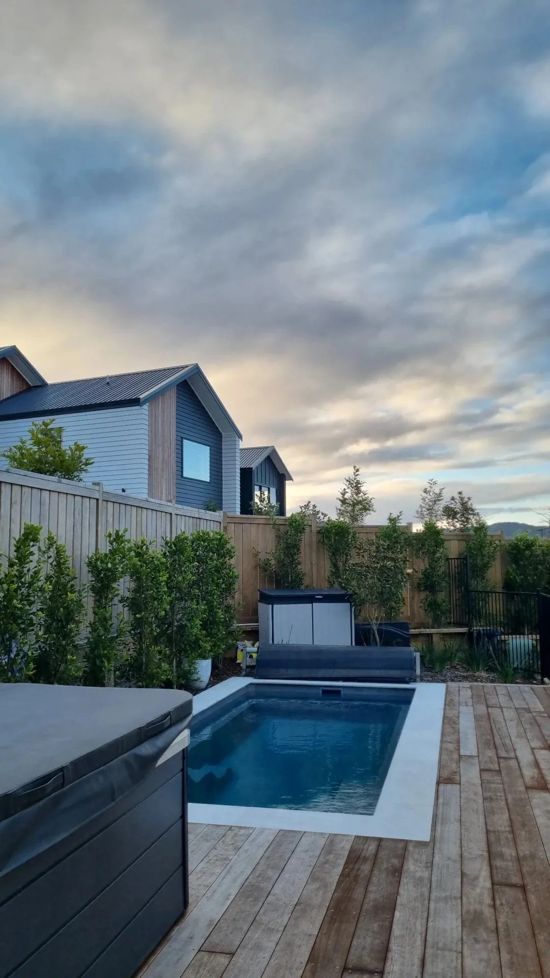 Backyard with a swimming pool, hot tub, deck, plants, wooden fence, and houses under a cloudy sky.