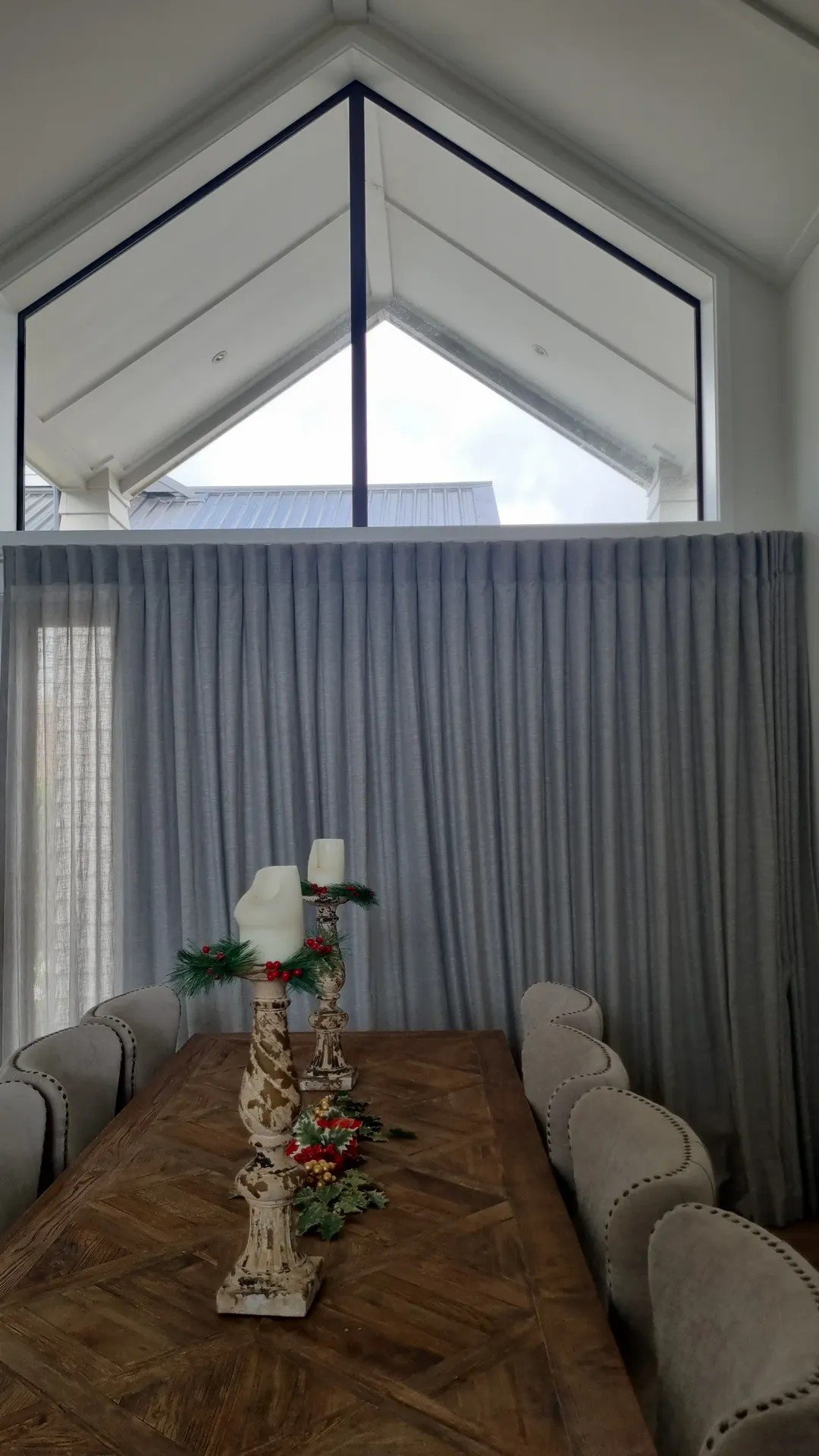 A dining room table with Christmas decorations, including candles, greenery, and red berries, surrounded by beige upholstered chairs. A large window with gray curtains is in the background, and the ceiling has a triangular design.