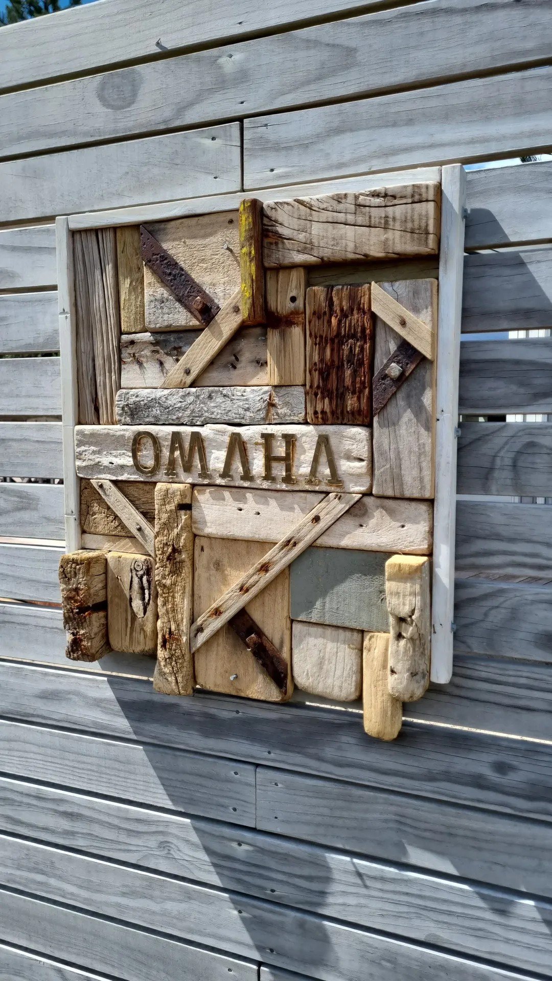 Wooden sign with Greek letters spelling OMHLA, mounted on a gray horizontal wooden wall.