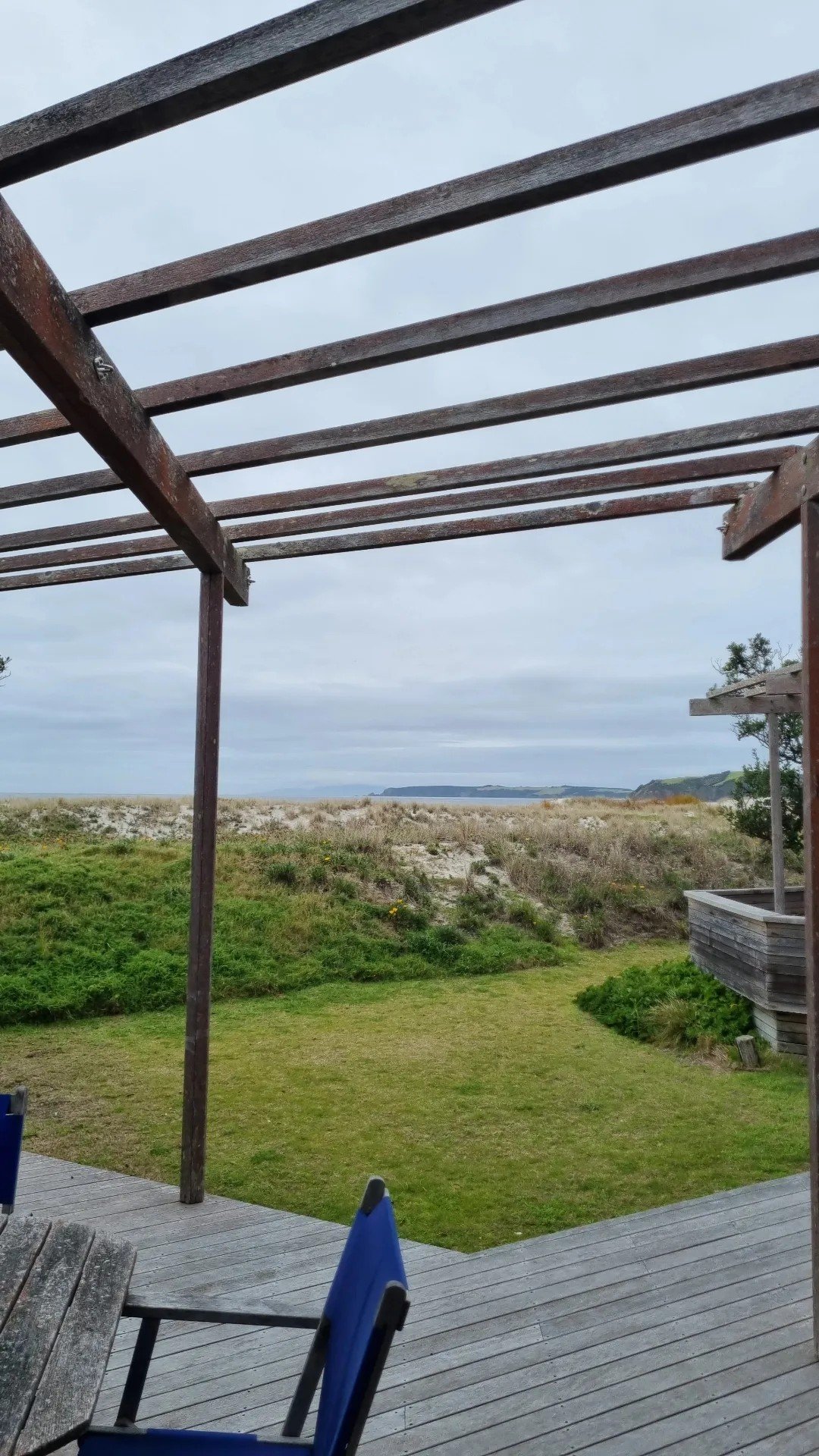View from a deck overlooking a grassy area, sand dunes, and the ocean under a cloudy sky, with part of a wooden pergola and blue chairs in the foreground.