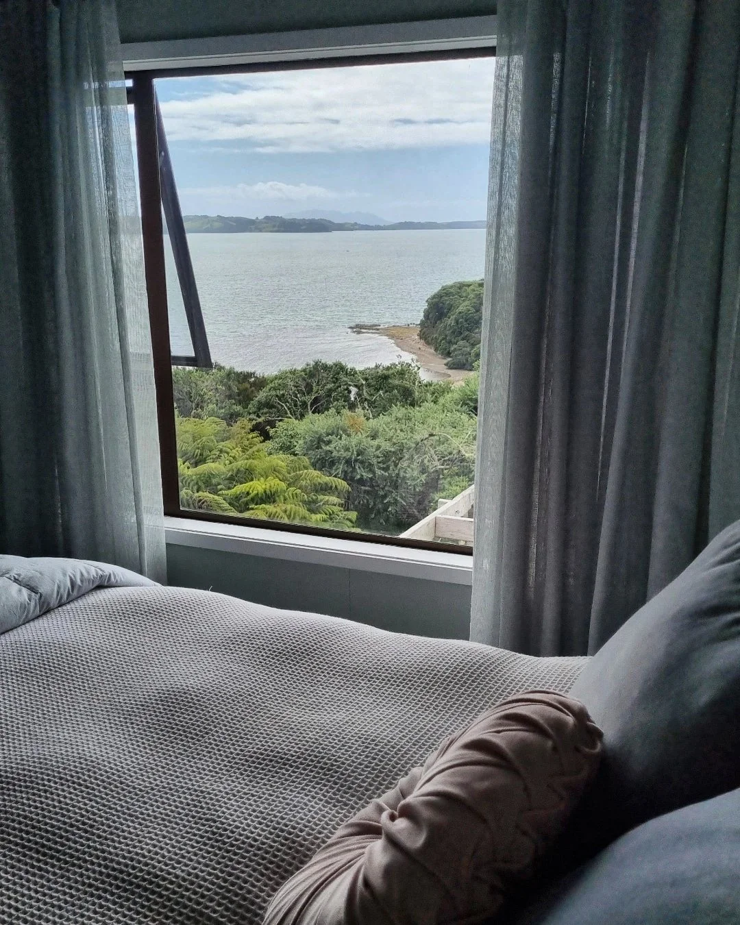 A bedroom with an ocean view through a large window, showing trees, water, and a cloudy sky.