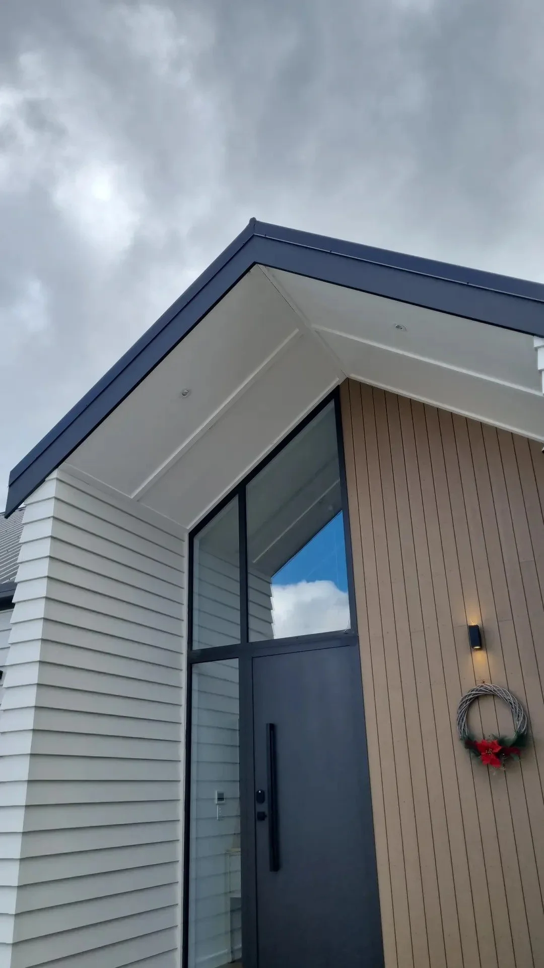 Modern house exterior showing a corner with white and wooden siding, large glass window, and a dark gray door with a wreath, under a cloudy sky.