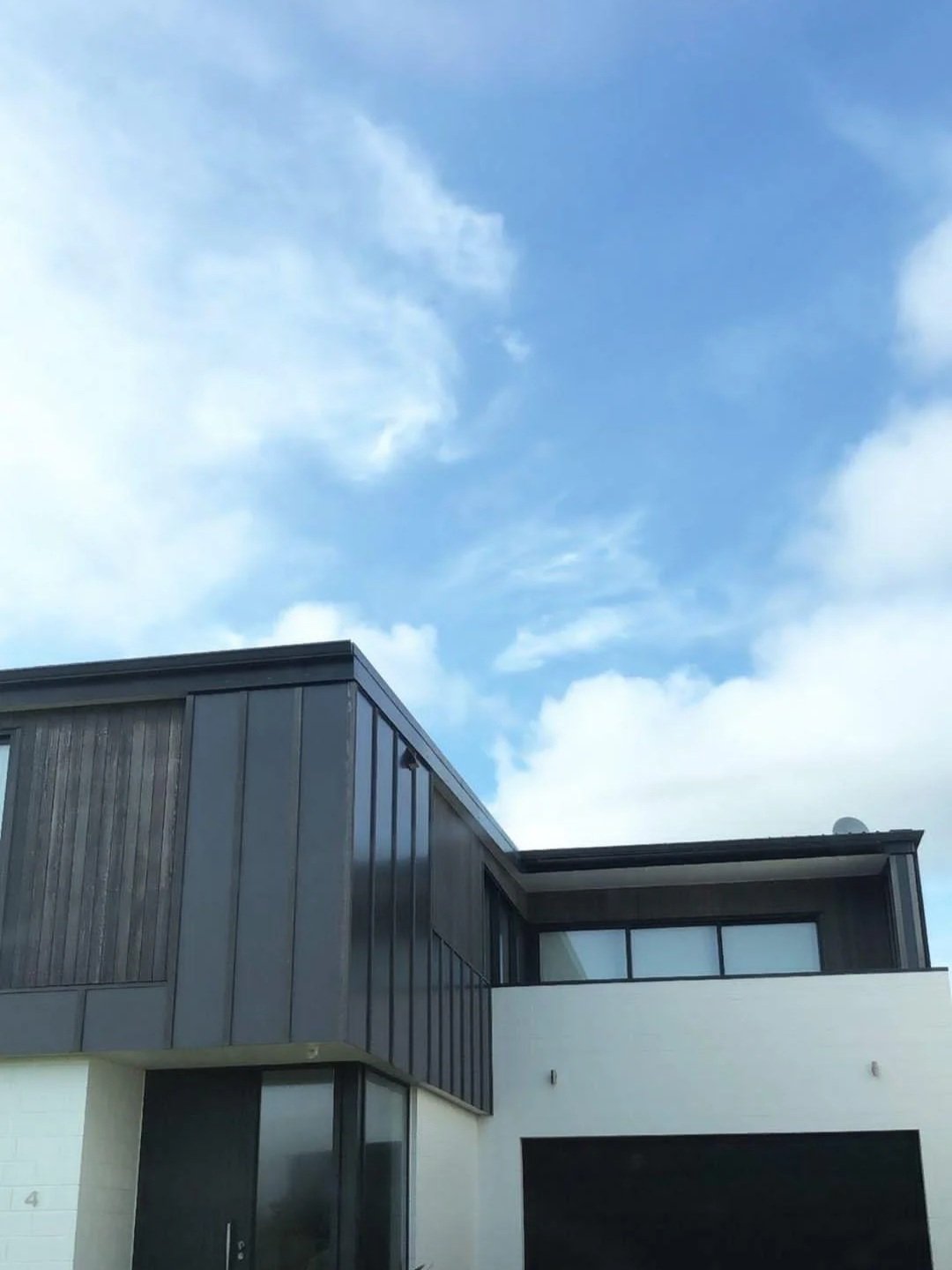 Modern house with black exterior panels and large windows, part of a multi-story building, under a partly cloudy blue sky.