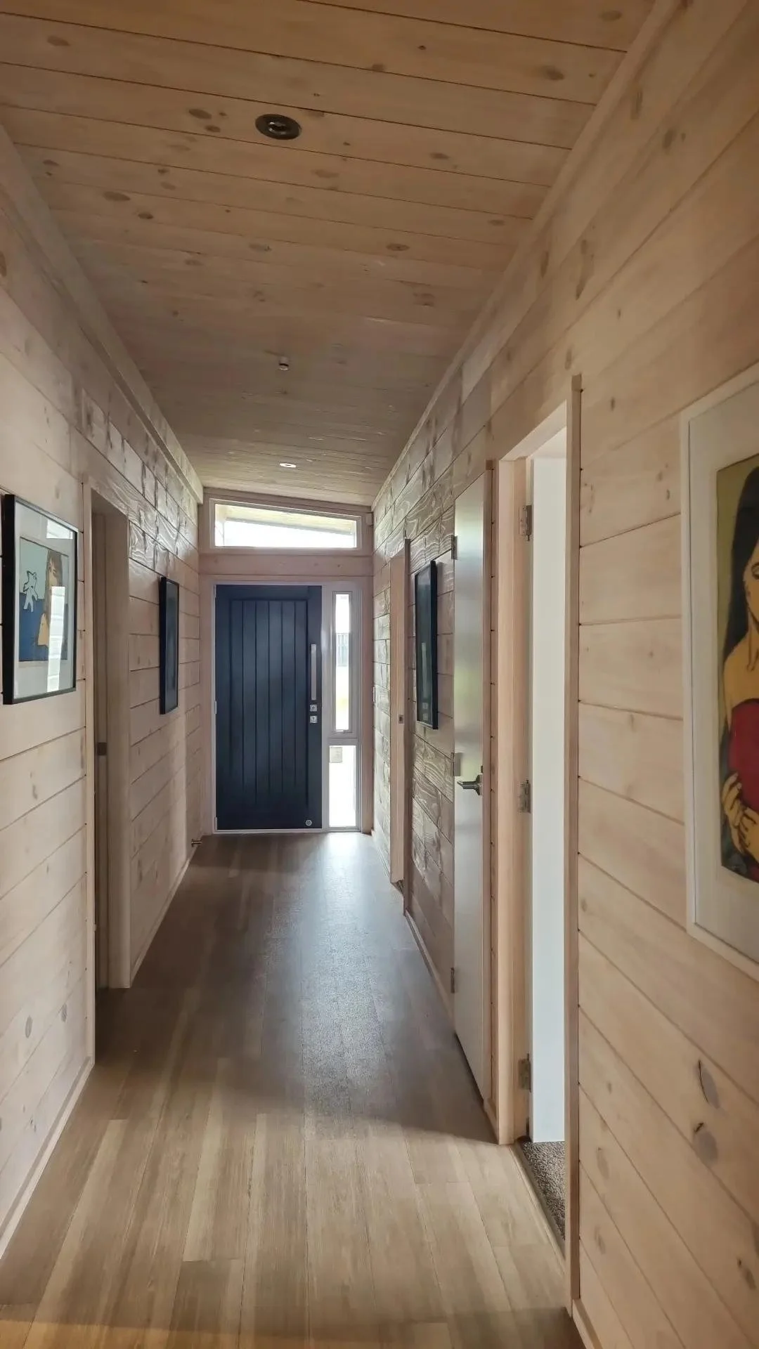 Wood-paneled hallway with framed artwork on the walls, a black front door with a vertical window. Light from a window above the door illuminates the space.