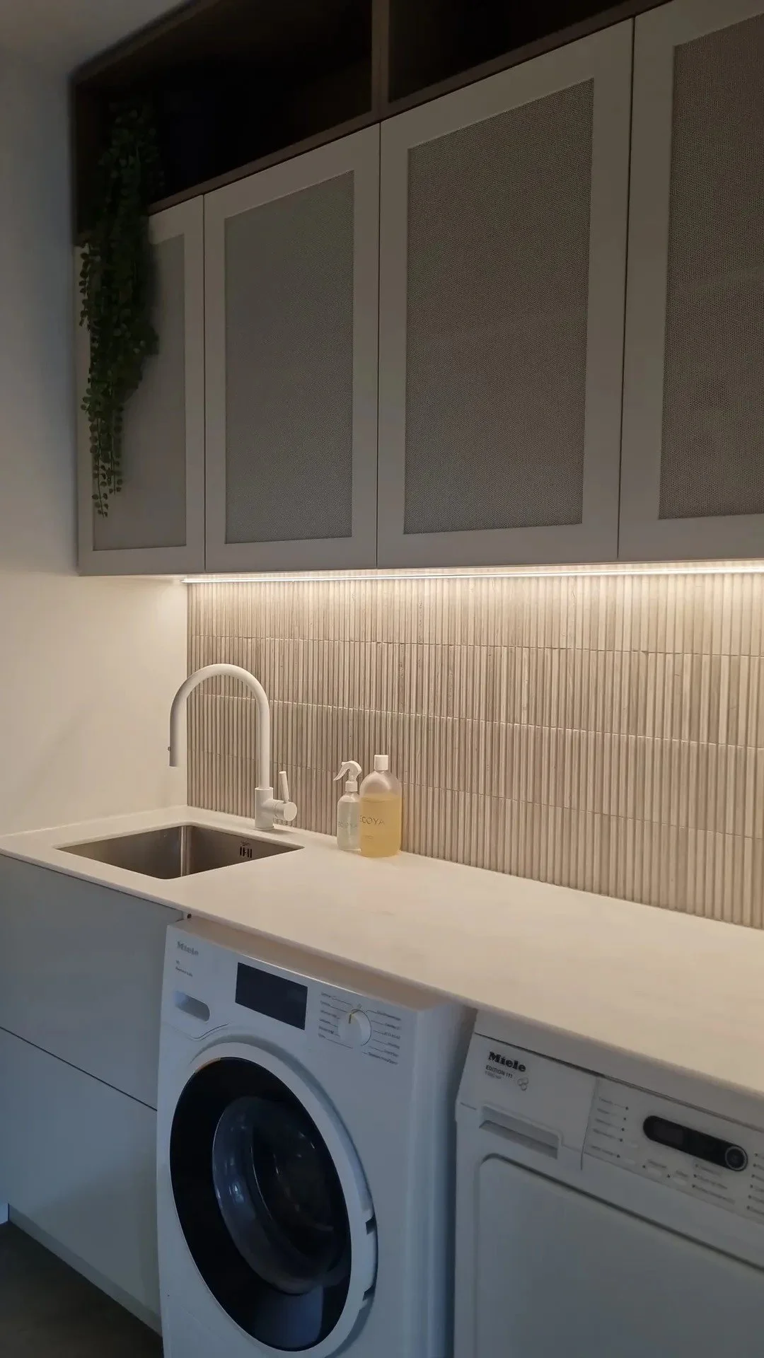 Modern laundry room with white washer and dryer, beige tiled backsplash, white countertop, and upper cabinets with mesh panels. There are cleaning supplies near the sink, which has a white faucet.