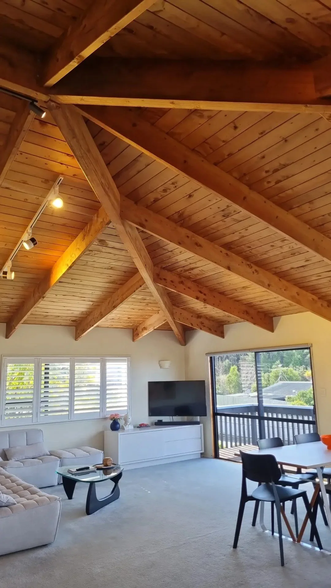 Living room with wooden vaulted ceiling, white sofa, black coffee table, TV on white cabinet, window with blinds, sliding glass door opening to balcony, and dining table with black chairs.