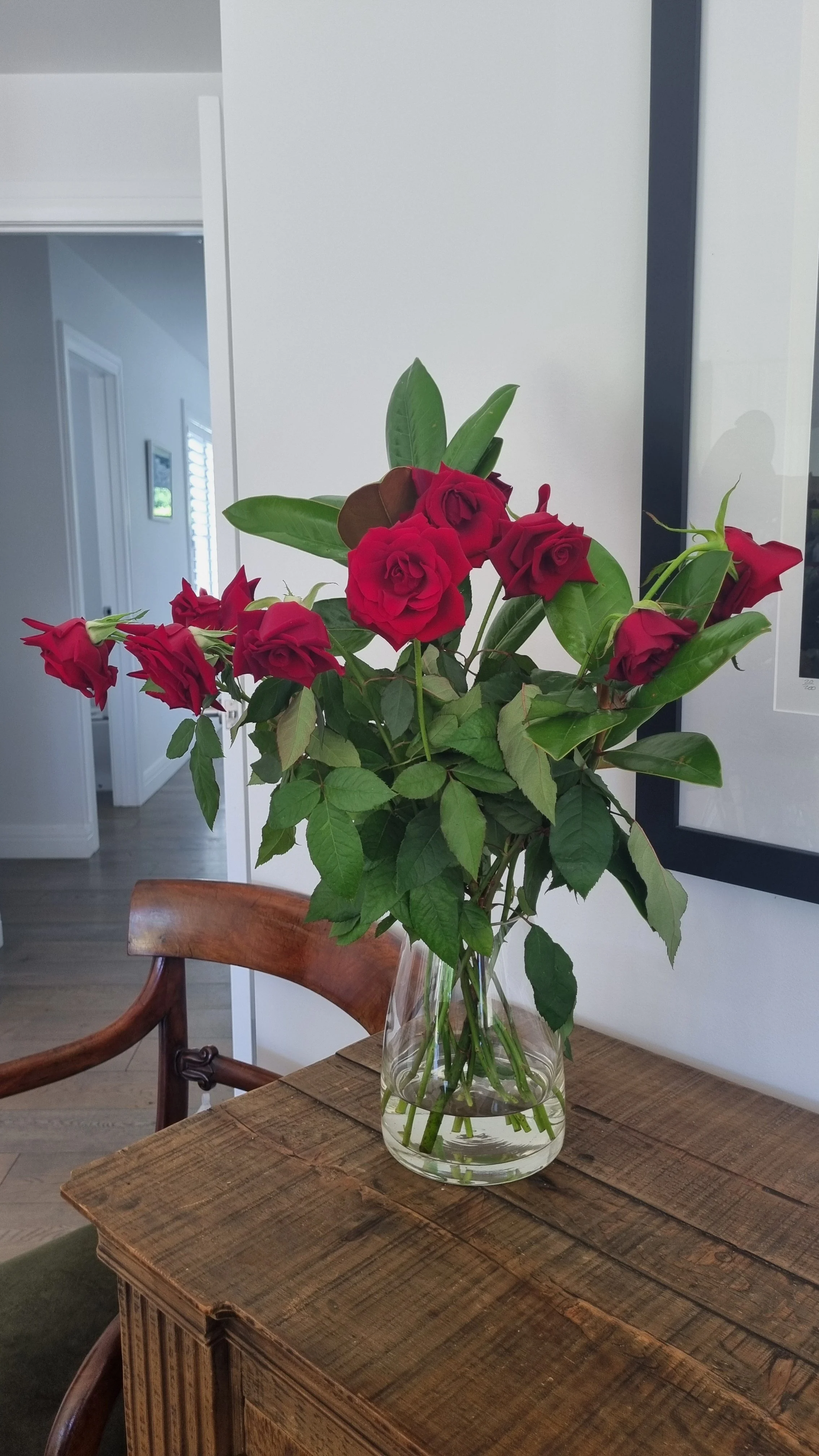 A bouquet of red roses in a clear glass vase on a rustic wooden table in a room with white walls and artwork.