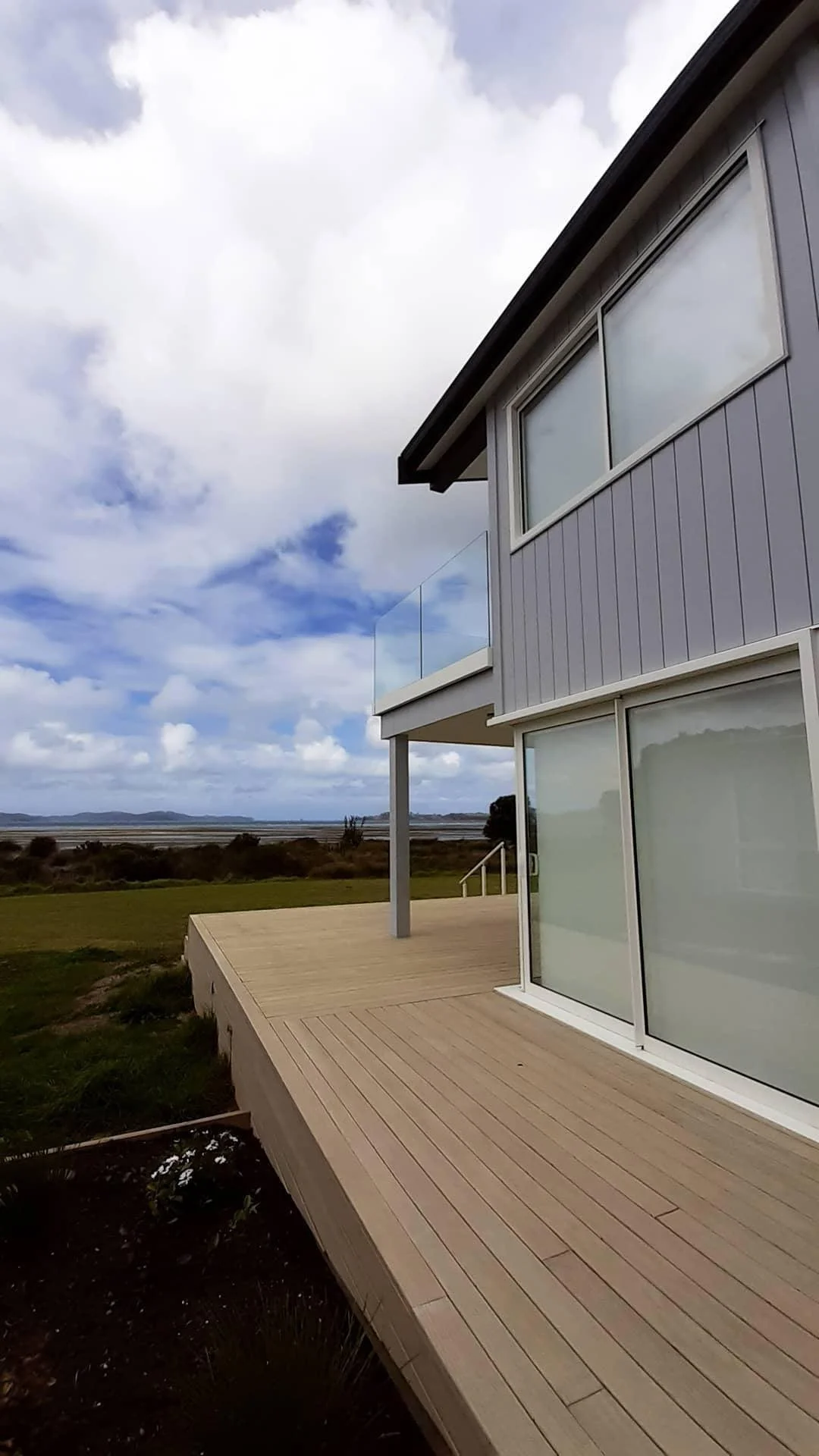 Modern two-story house with large glass windows and a wooden deck overlooking open fields and a cloudy sky.