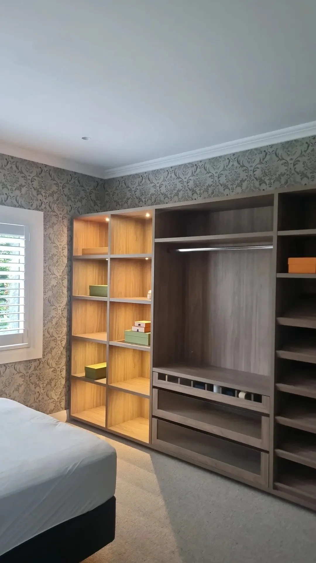 Room with built-in shelving, patterned wallpaper, window with white blinds, bed with white mattress, and beige carpet.
