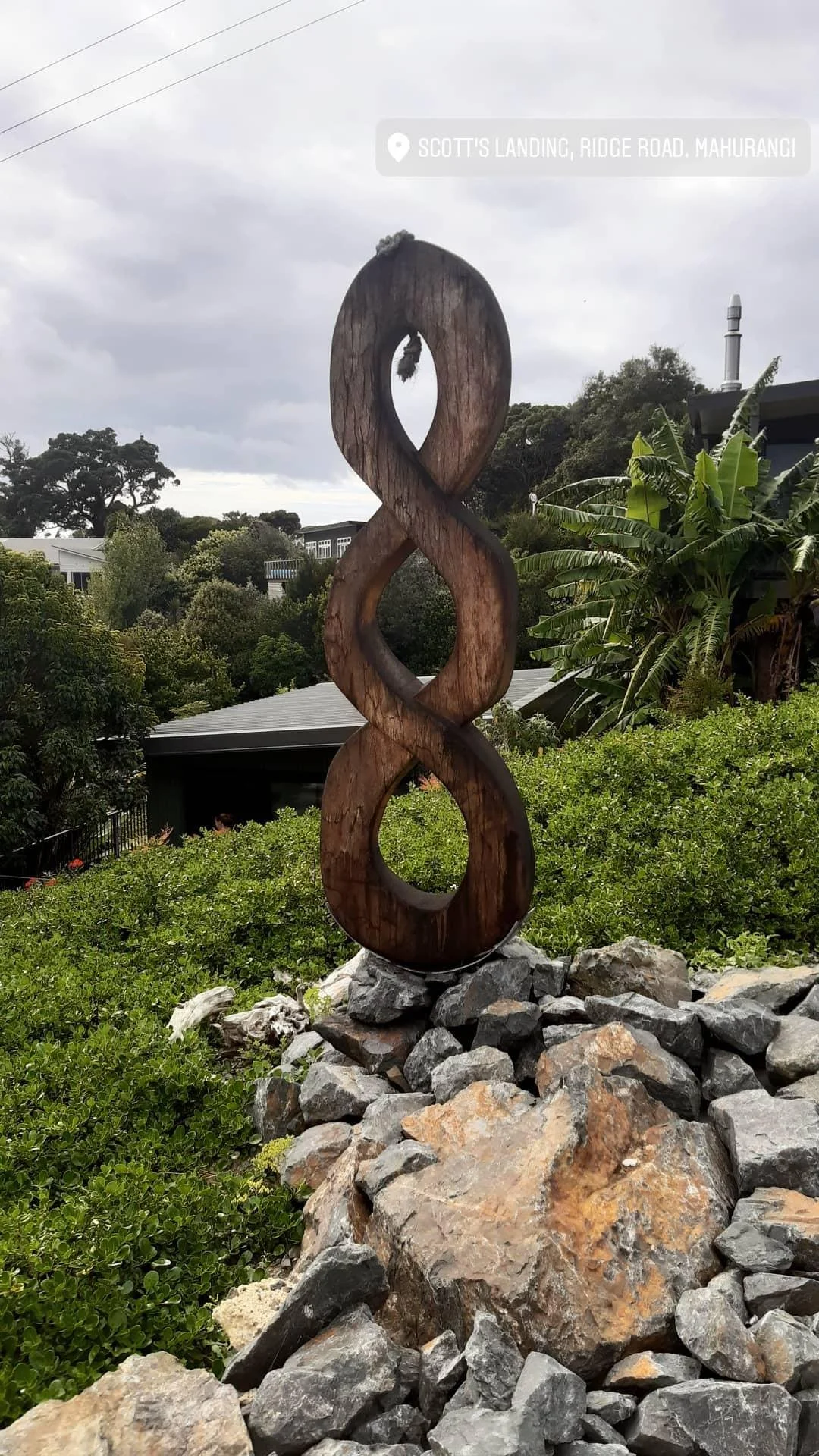 Sculpture of a wooden infinity symbol on a rocky base, with greenery and trees in the background, and cloudy sky above.
