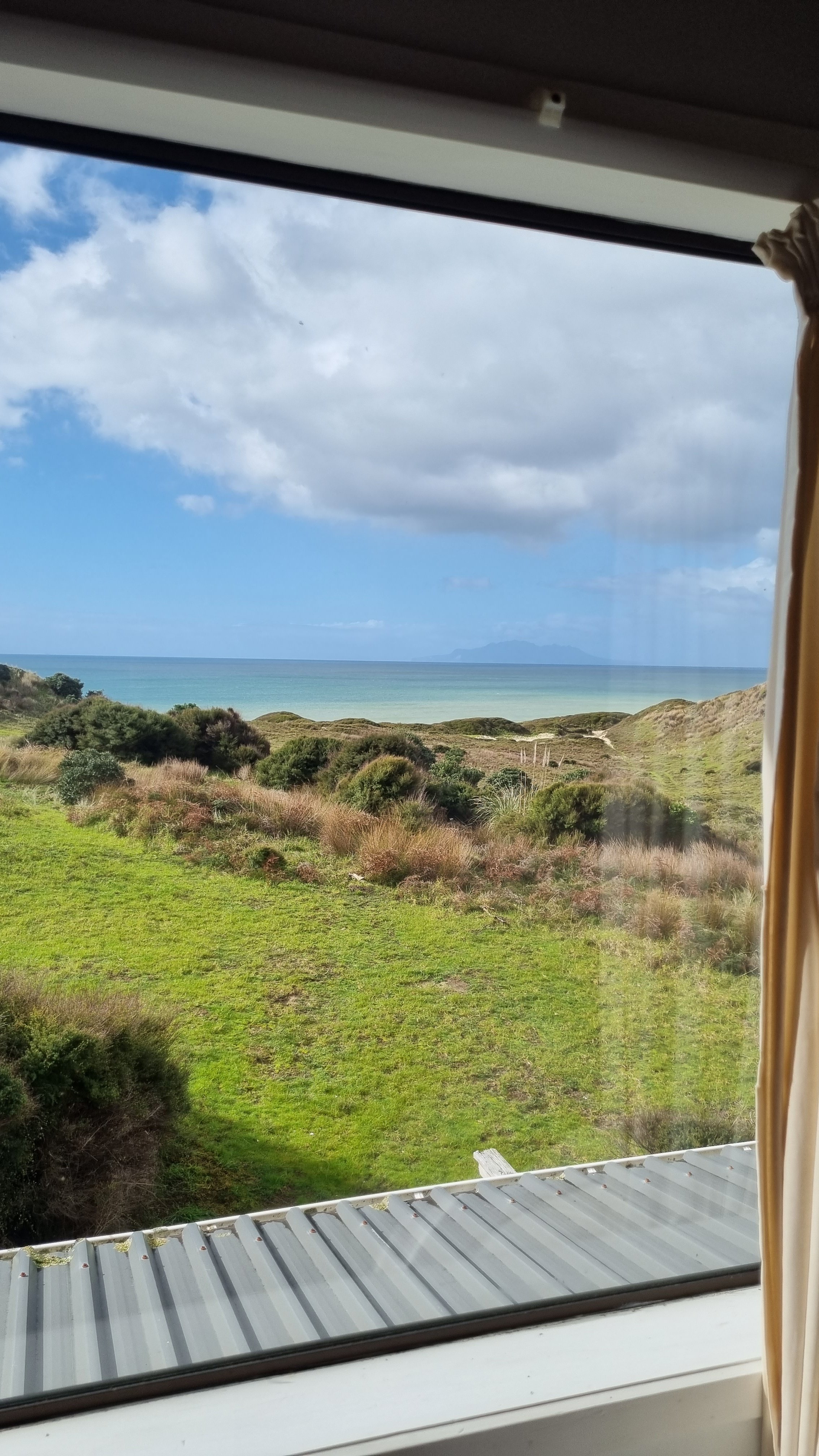 View through a window showing a grassy hillside, bushes, and the ocean with a distant mountain range under a partly cloudy sky.