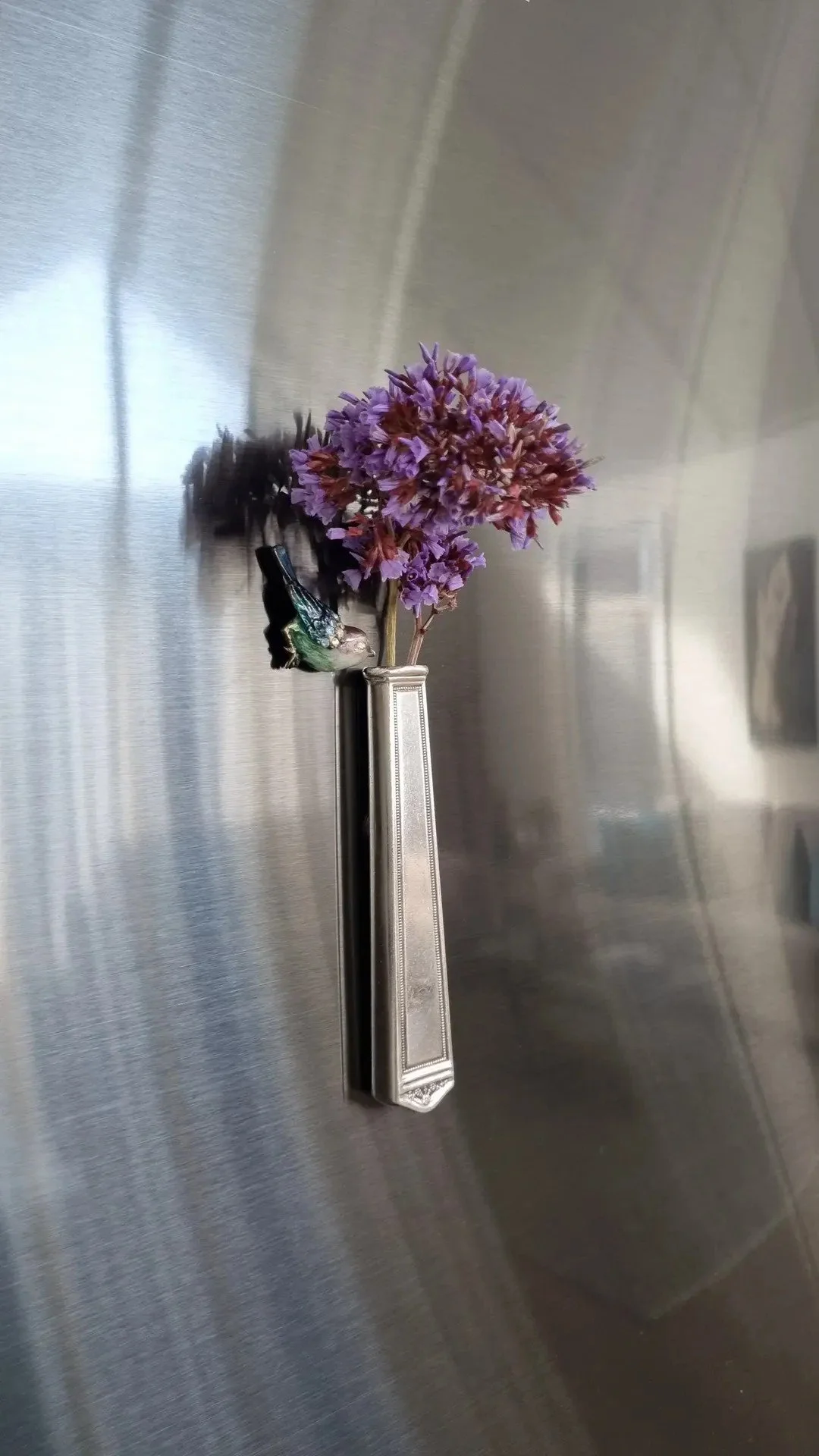 A small bunch of purple flowers in a silver vase on a stainless steel refrigerator door, with a decorative bird figure attached to the vase.
