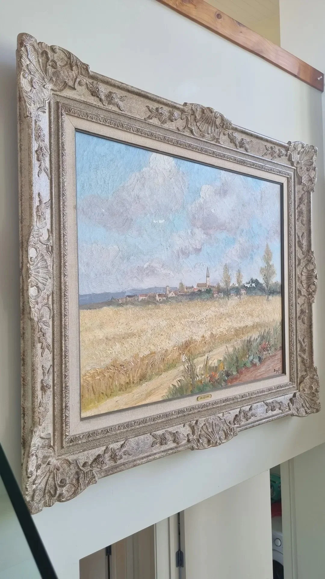 Landscape painting of a rural scene with golden fields, a small village with church steeple in the distance, and blue sky with clouds, framed in an ornate silver-colored frame.