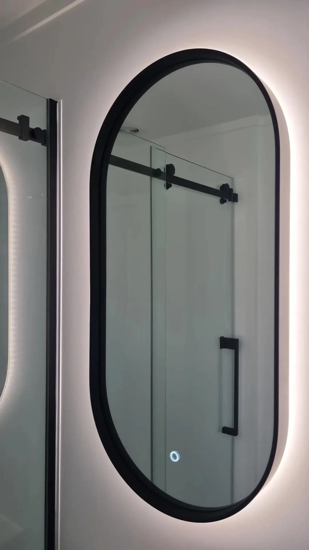 An oval mirror with black frame mounted on a light-colored wall in a modern bathroom, reflecting a shower door and handle.