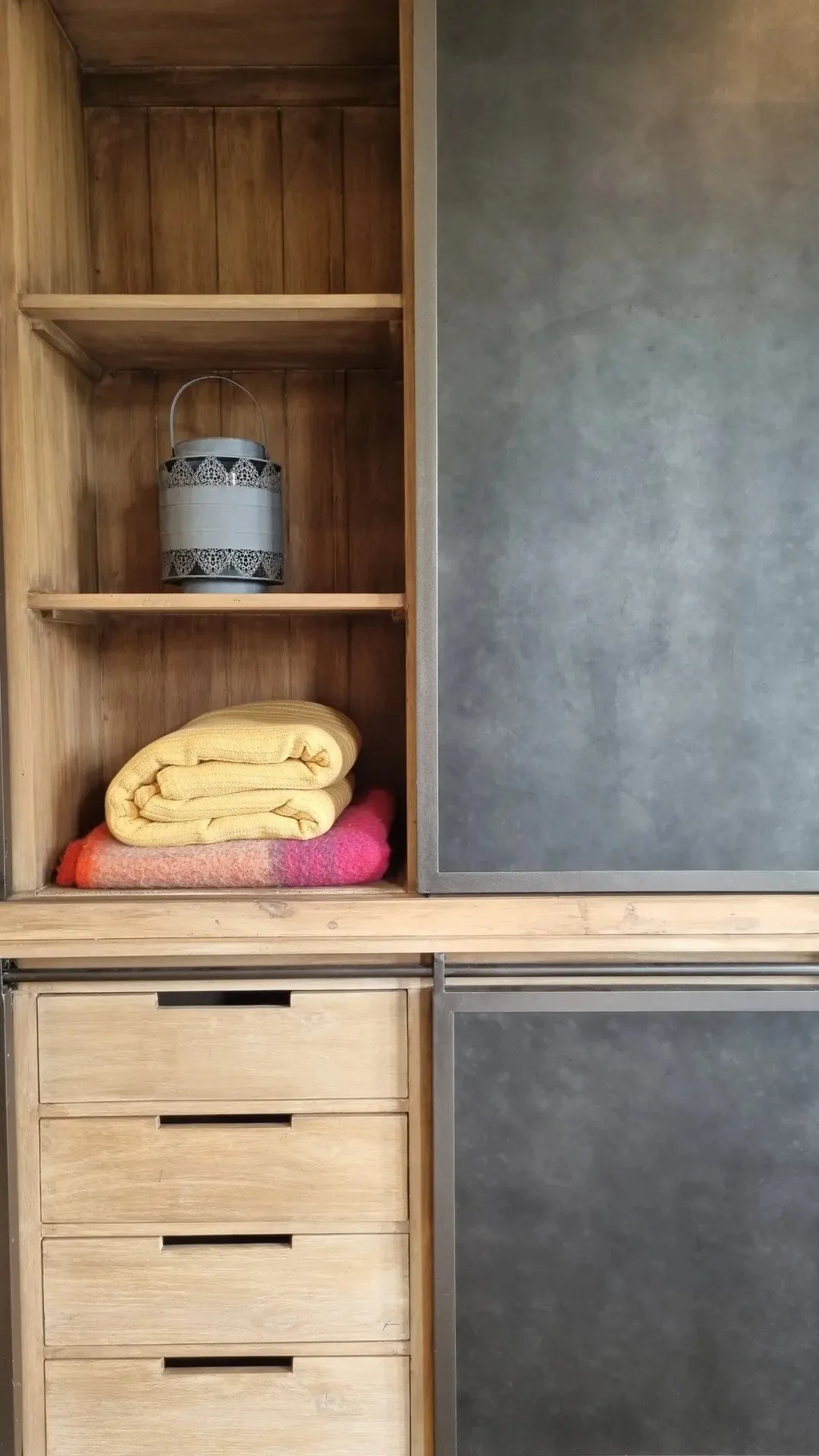 Open wooden cabinet with three yellow towels and one pink towel on the bottom shelf, and a decorative metal container on the middle shelf.