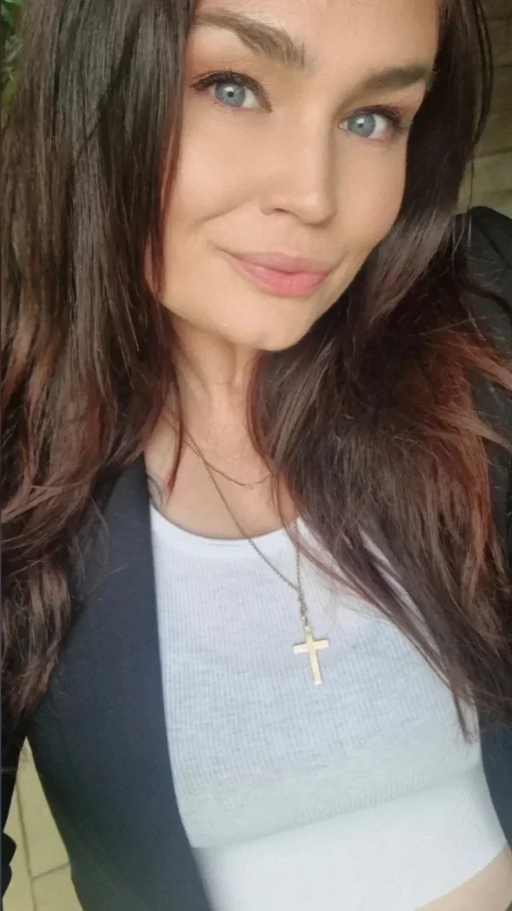 Close-up selfie of a woman with long brown hair, blue eyes, and light skin, wearing a white top, black jacket, and a gold cross necklace.