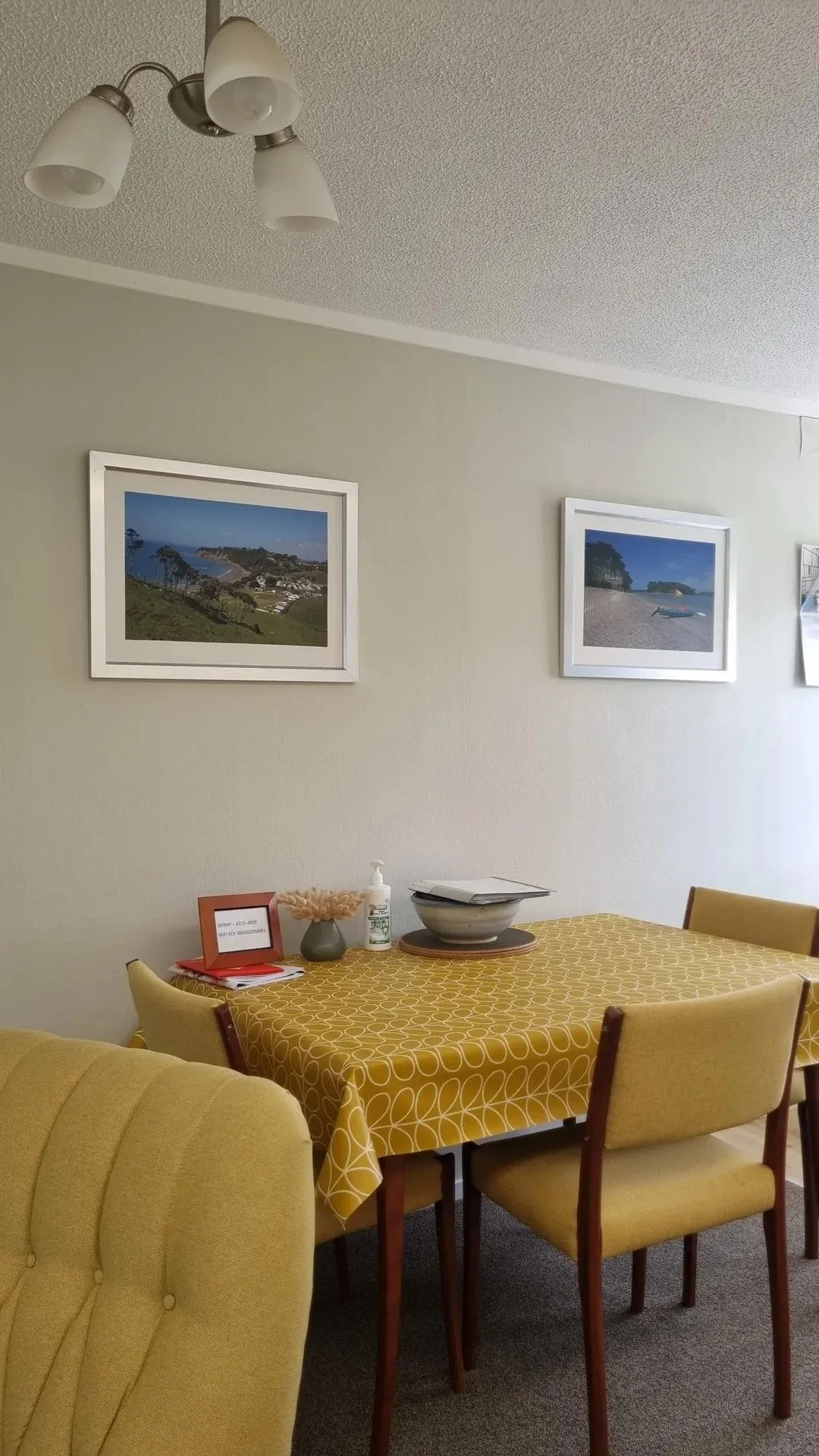Interior of a dining room with a yellow tablecloth, four yellow chairs, framed landscape photos on the wall, and a ceiling light fixture.