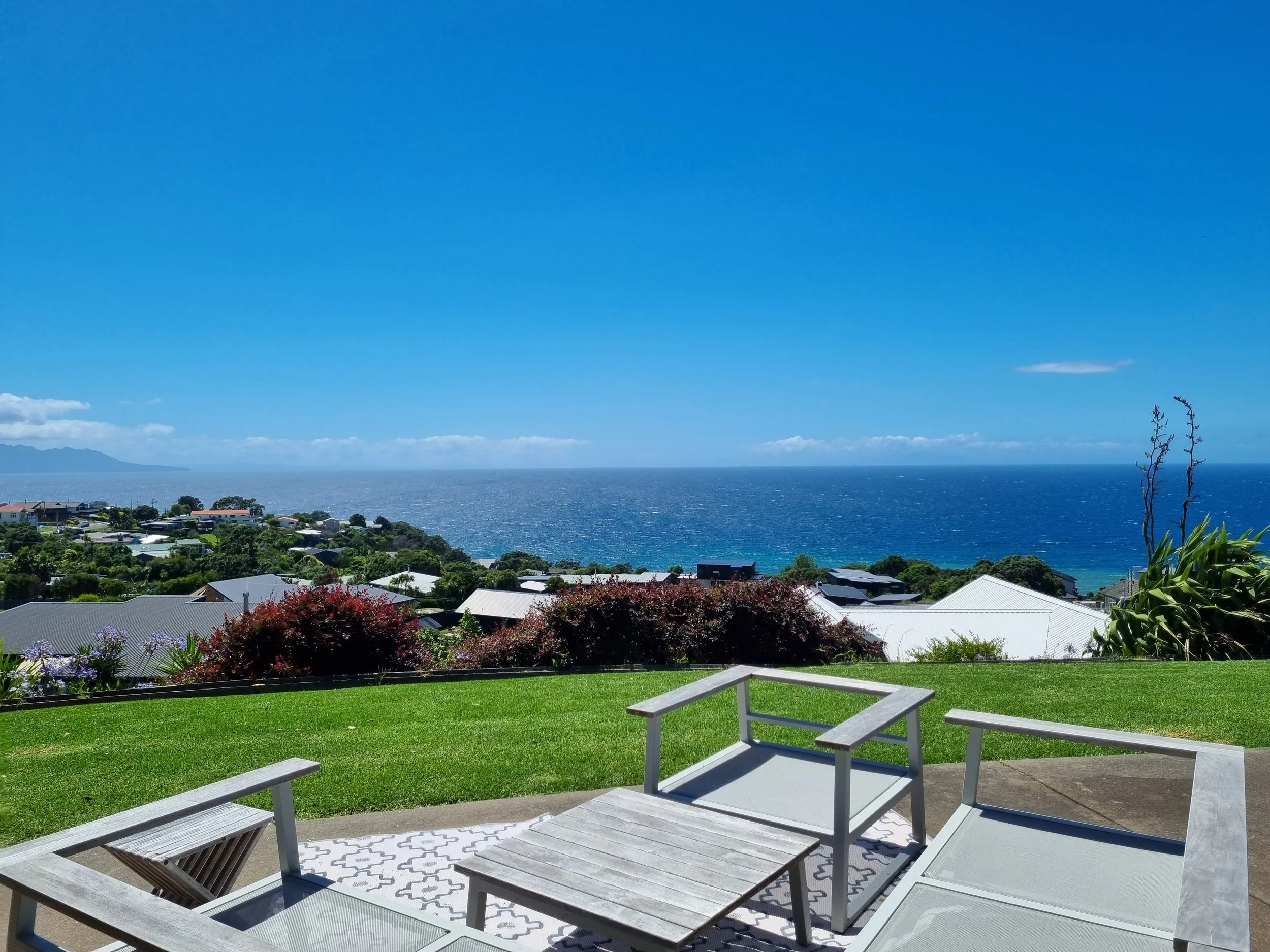 A backyard with a wooden deck, white outdoor furniture including a table and chairs, green grass, flowering bushes, and a view of the ocean with a distant mountain range under a clear blue sky.