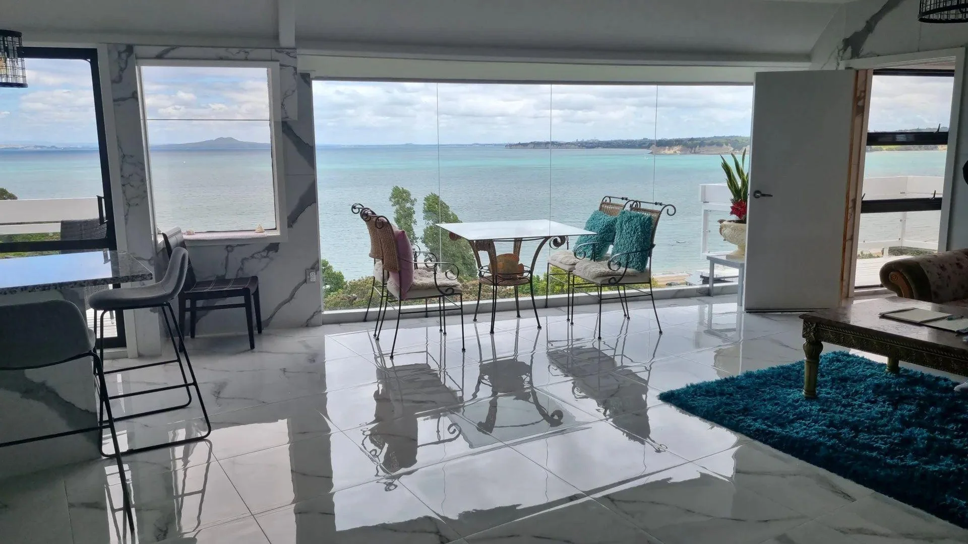 Living room with large glass windows overlooking a body of water, with a dining table and chairs, a floral sofa, a blue rug, potted plants, and marble flooring.