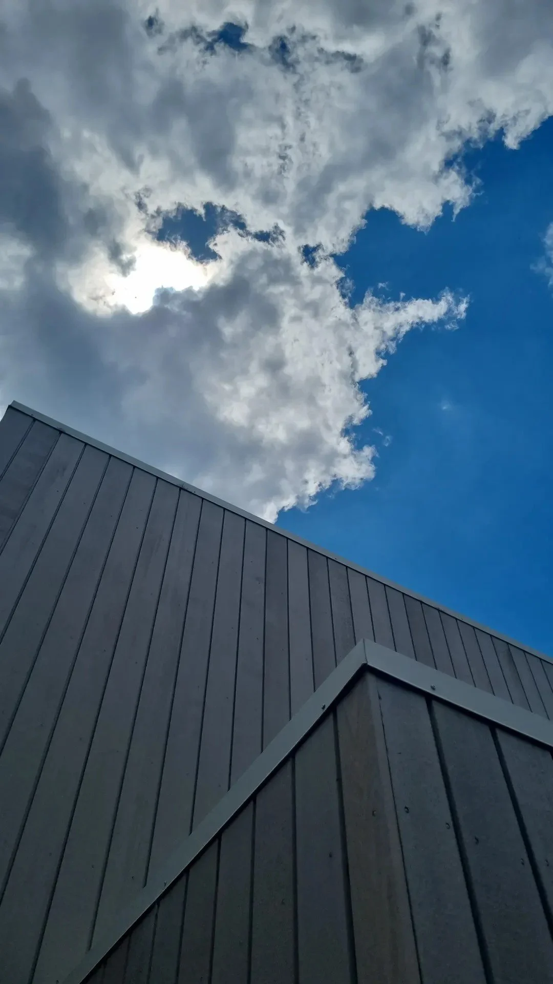 Part of a modern gray building with vertical siding against a background of partly cloudy sky with blue and white clouds.