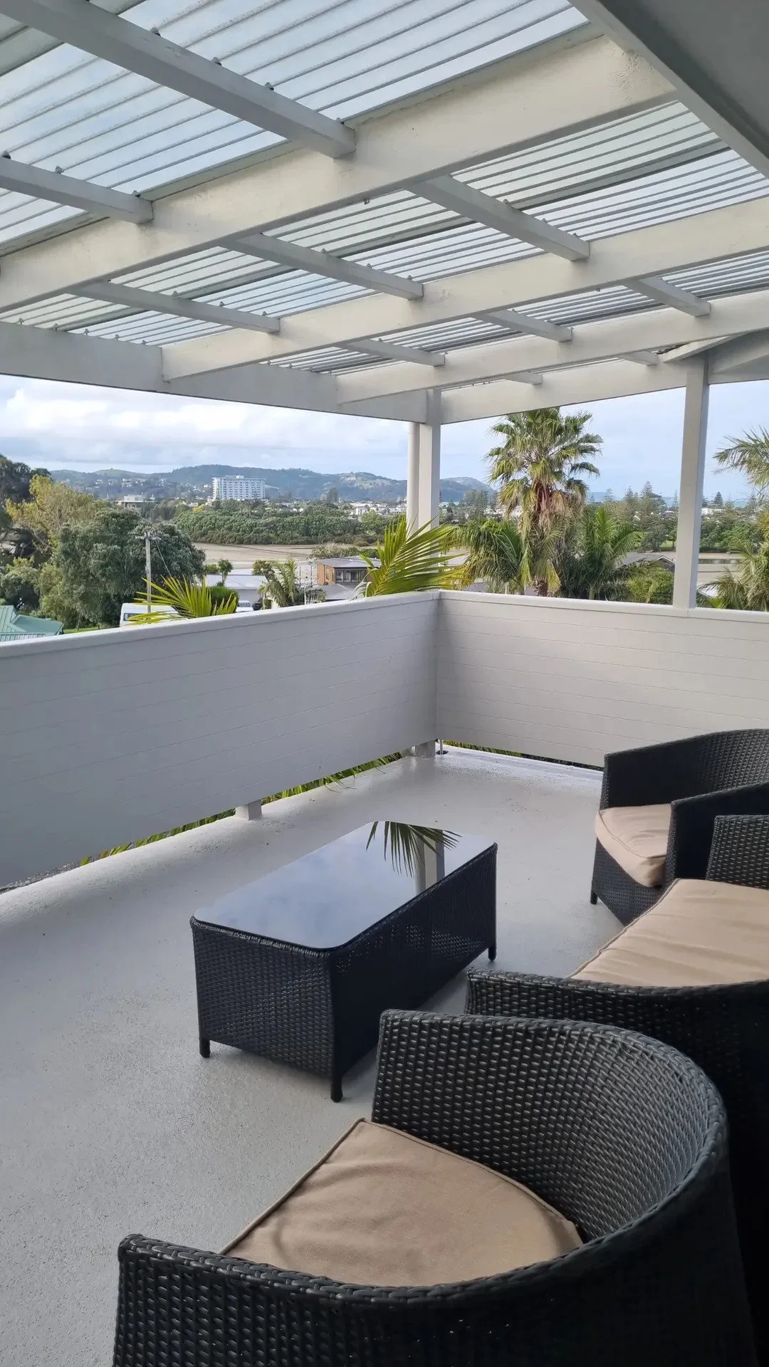 Balcony with wicker furniture, a glass-top table, and potted palm plants, overlooking a landscape with trees, hills, and buildings under a partly cloudy sky.