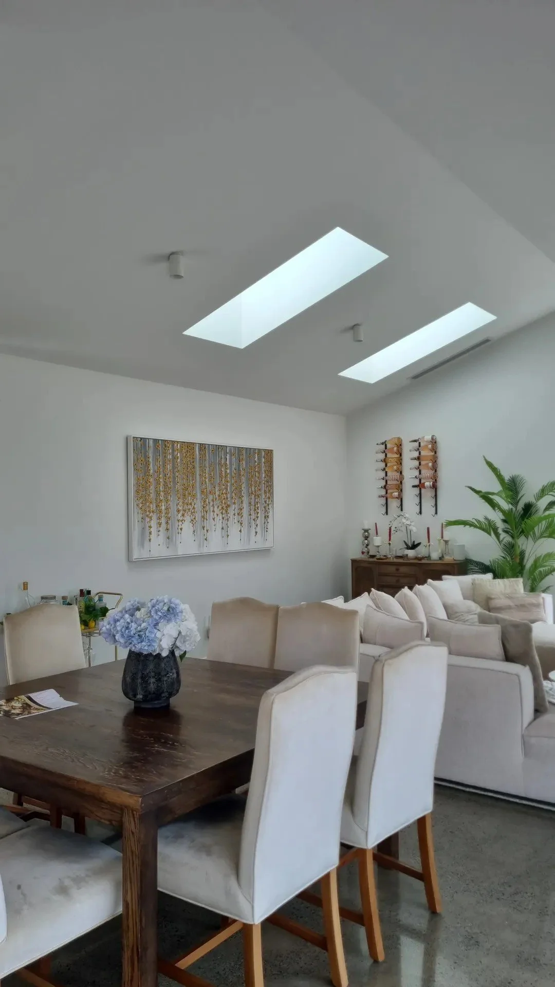 Bright living room with skylights, a wooden dining table with cream chairs, a beige sofa, artwork on the wall, a large potted plant, and decorative shelves with wine bottles.