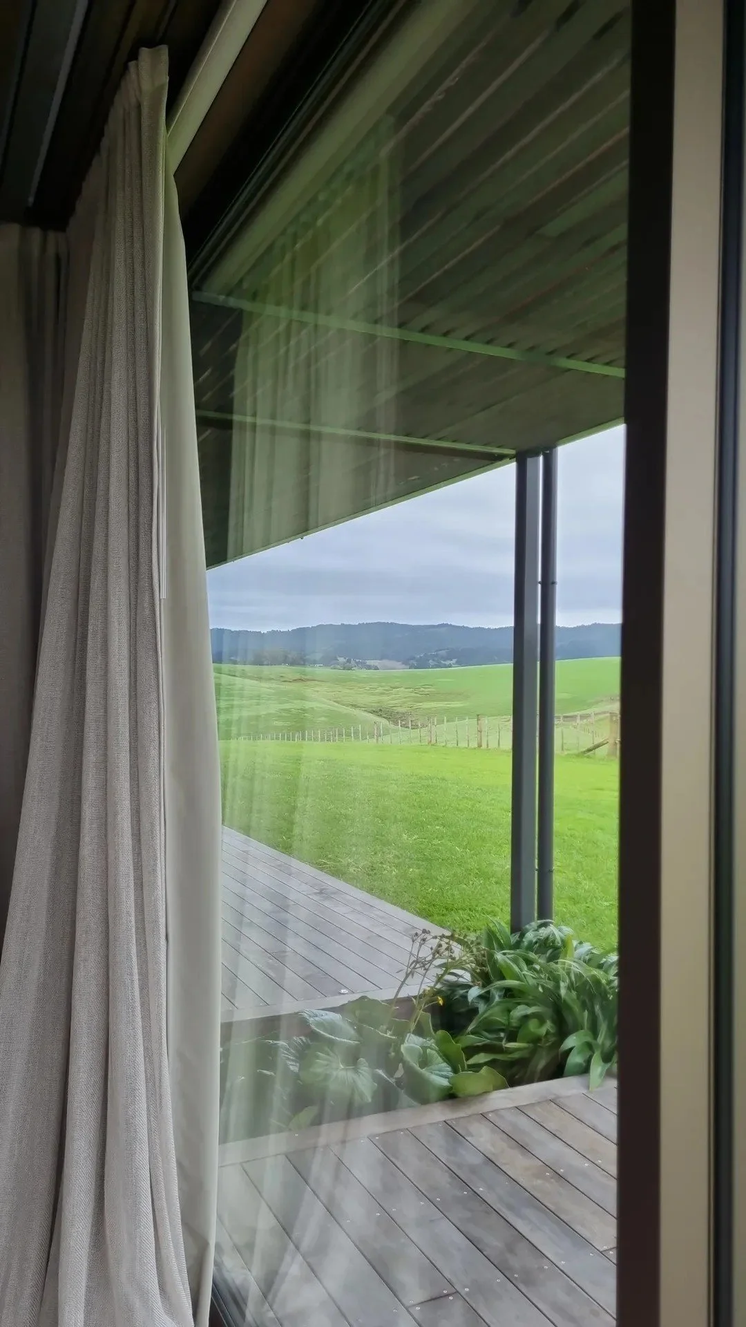 View through a large glass window showing a green yard, a wooden deck, and rolling hills in the distance under a cloudy sky.