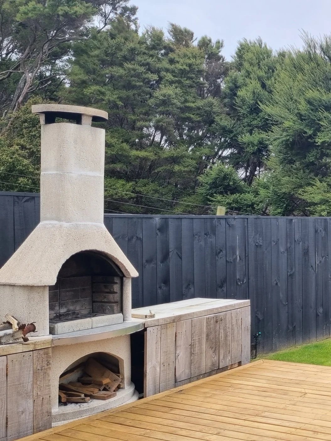 An outdoor fireplace and pizza oven setup with a wooden counter and a fenced yard, trees in the background.