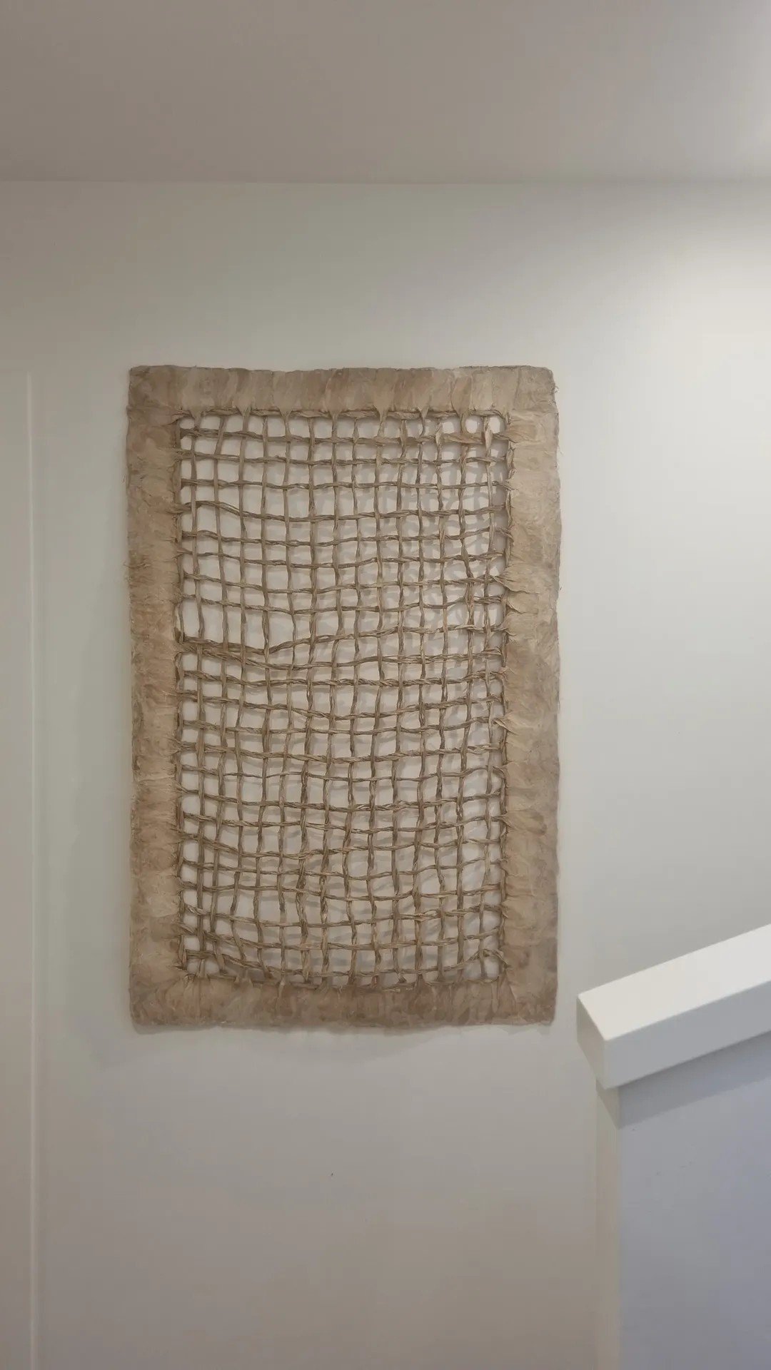A beige woven mat with a netted pattern hangs on a white wall in a minimalist interior space.