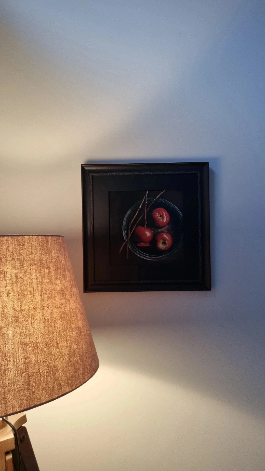 A framed still life painting of three red apples with two small sticks, hanging on a white wall near a beige lampshade.