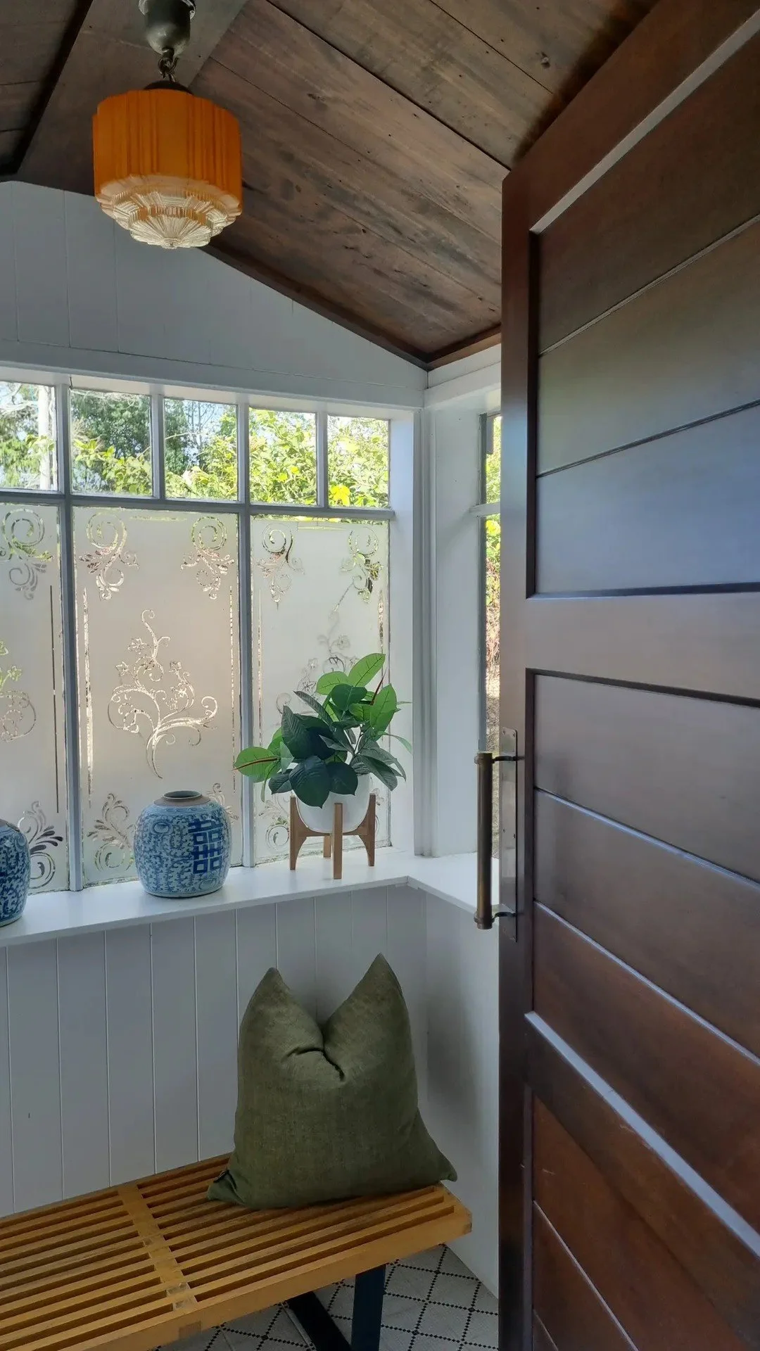 View into a cozy entryway with wooden ceiling, hanging orange lamp, decorative frosted glass window with floral patterns, potted green plant on windowsill, blue and white ceramics, olive green cushion on a wooden bench, and an open wooden door.