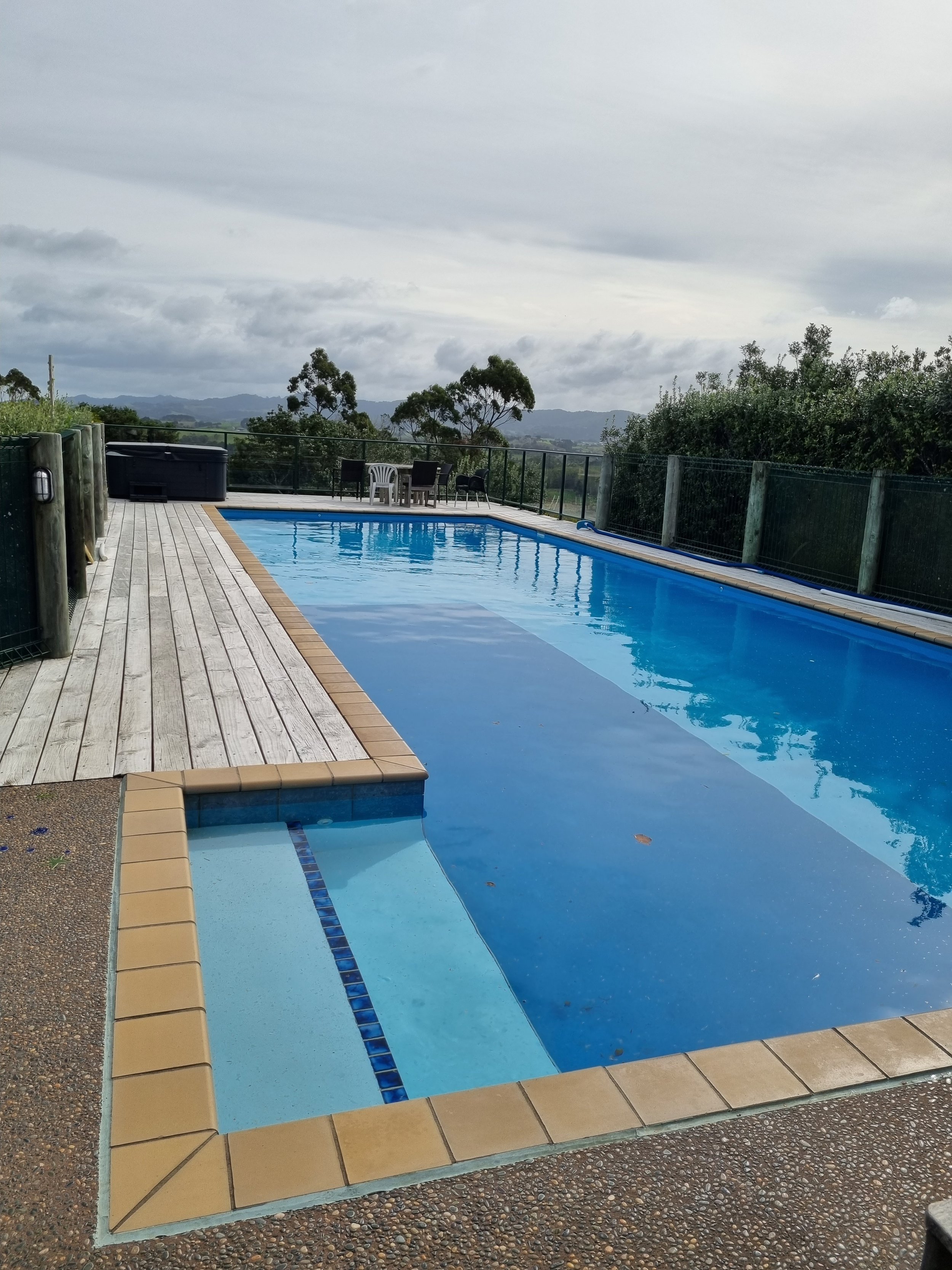 An outdoor swimming pool with blue water, surrounded by a wooden deck and beige tiles. In the background, there are trees, a cloudy sky, and a distant view of hills.