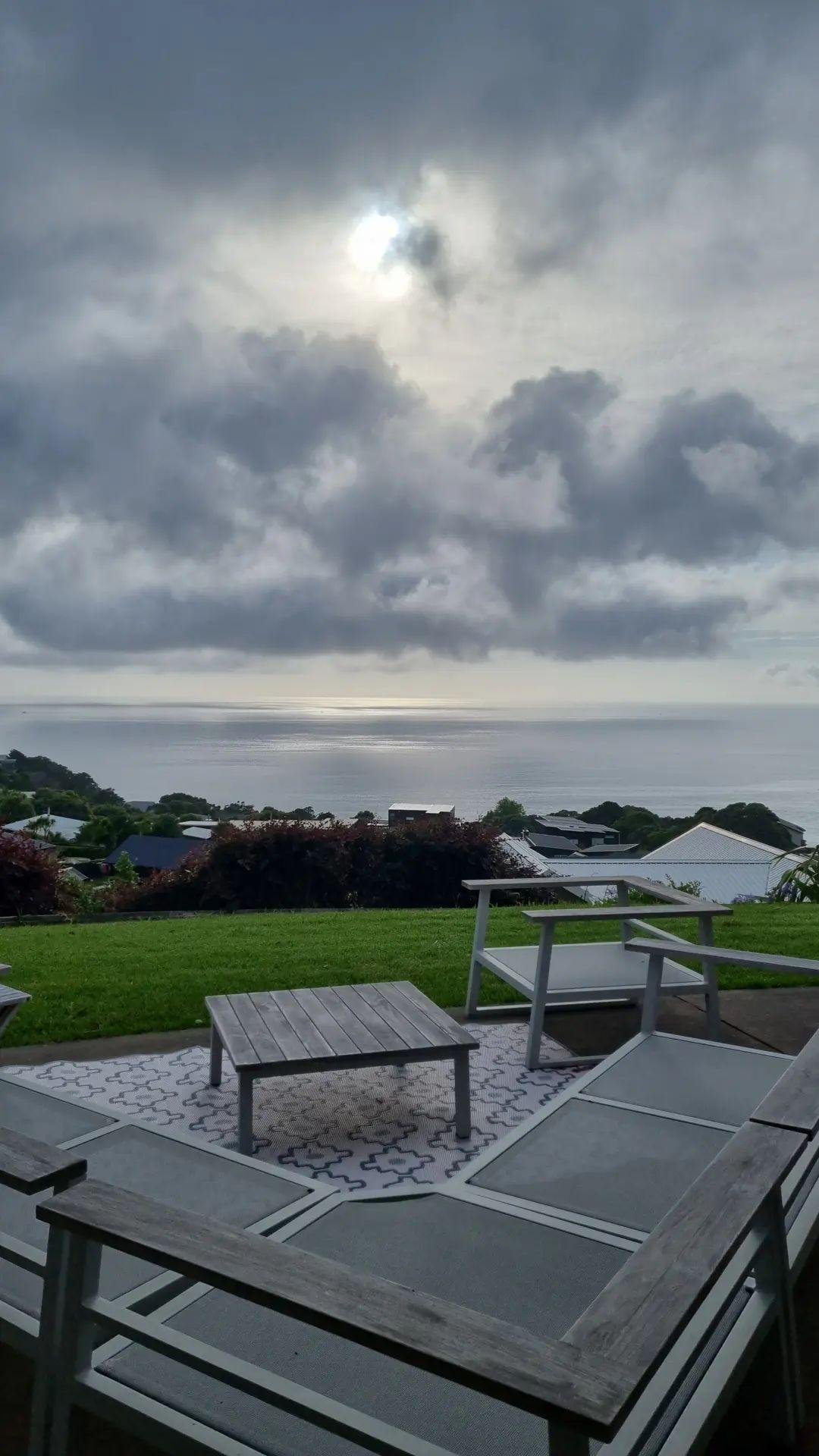 View from a patio showing outdoor chairs and tables overlooking a calm body of water under a cloudy sky with some sunlight breaking through.