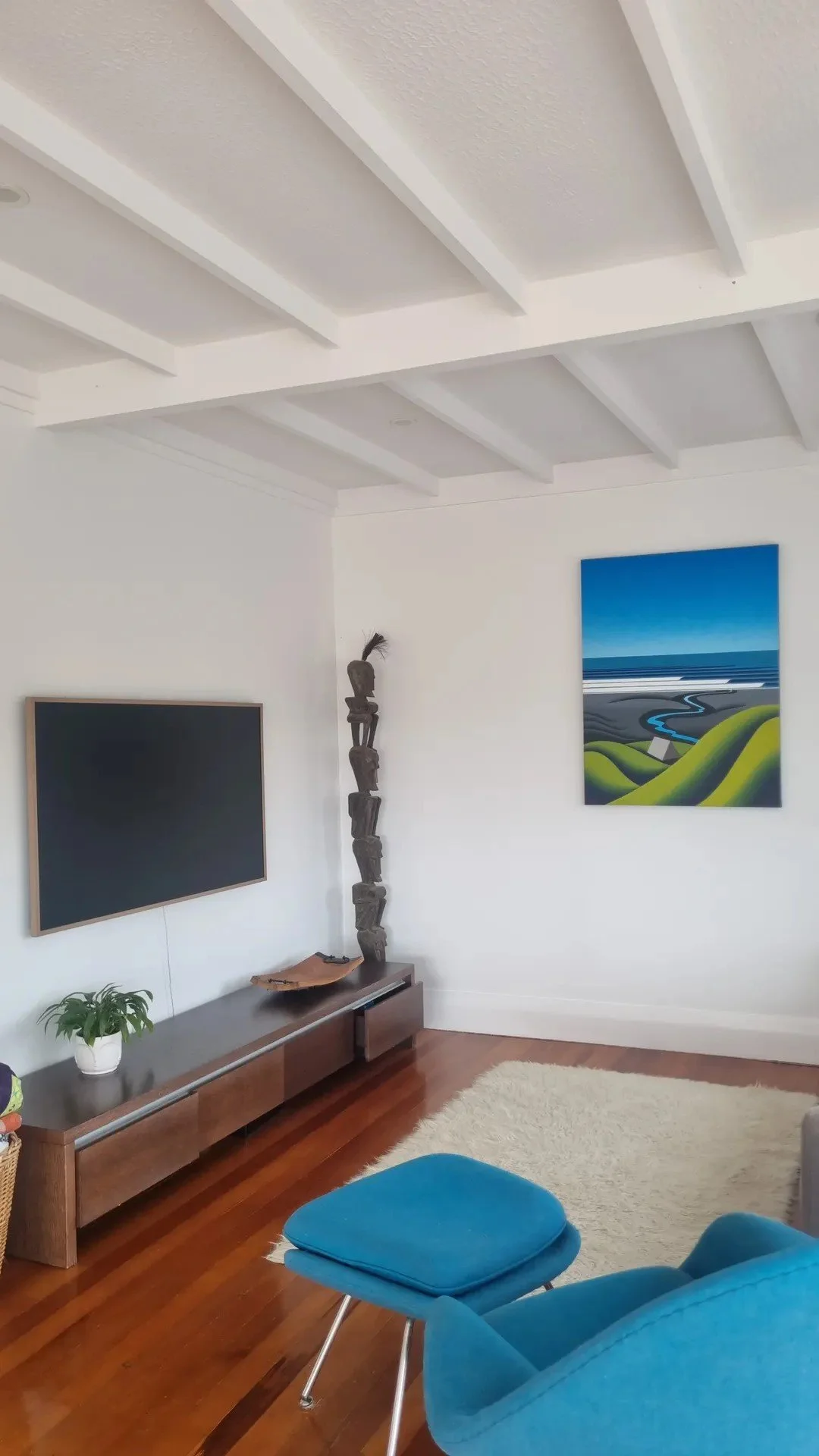 Living room with white ceiling featuring exposed beams, a wall-mounted flat-screen TV, a small wooden console table with a plant, a unique sculpture made of stacked rocks, a colorful landscape painting, a beige rug, and a blue armchair with a matchin