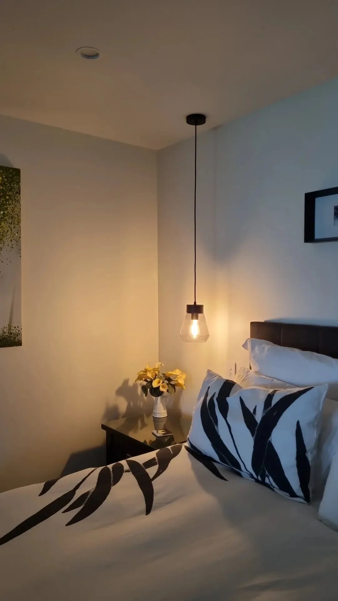 A bedroom with a bed, black and white patterned bedding, and pillows. There is a nightstand with a vase of yellow flowers, a small electronic device, and a hanging pendant light. Part of a framed picture is visible on the wall.