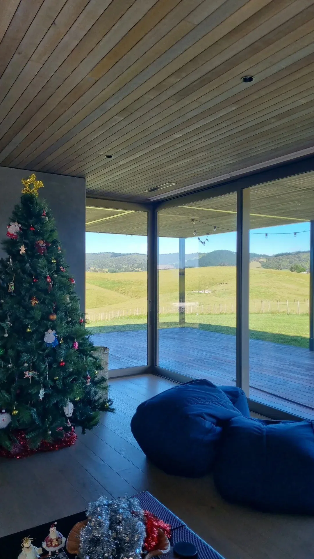 A living room with a decorated Christmas tree, large glass windows showing green hills, and a mountain range in the distance. There is a bean bag chair and a table with holiday decorations.