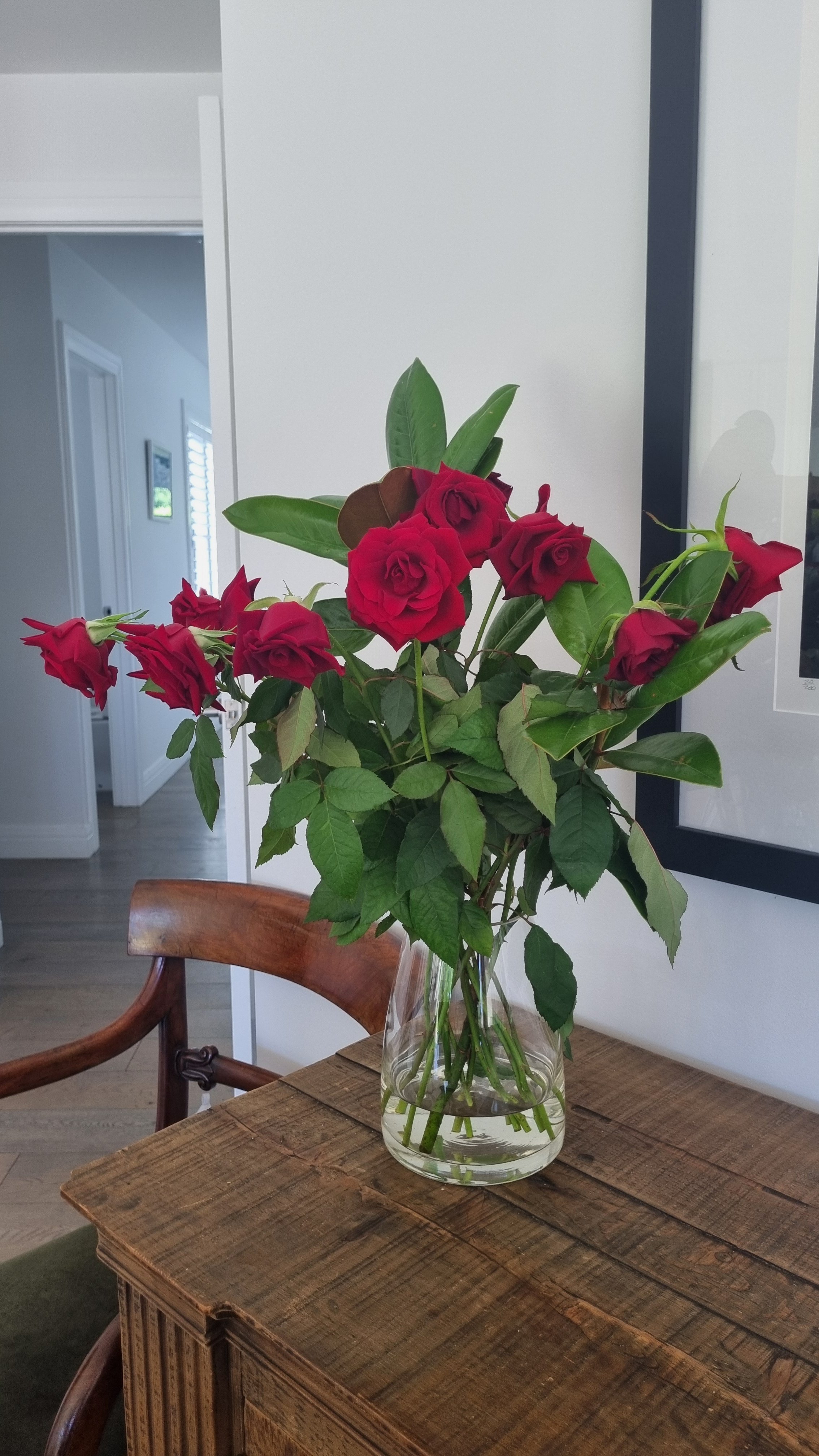 A bouquet of red roses and green leaves in a clear glass vase on a wooden table.
