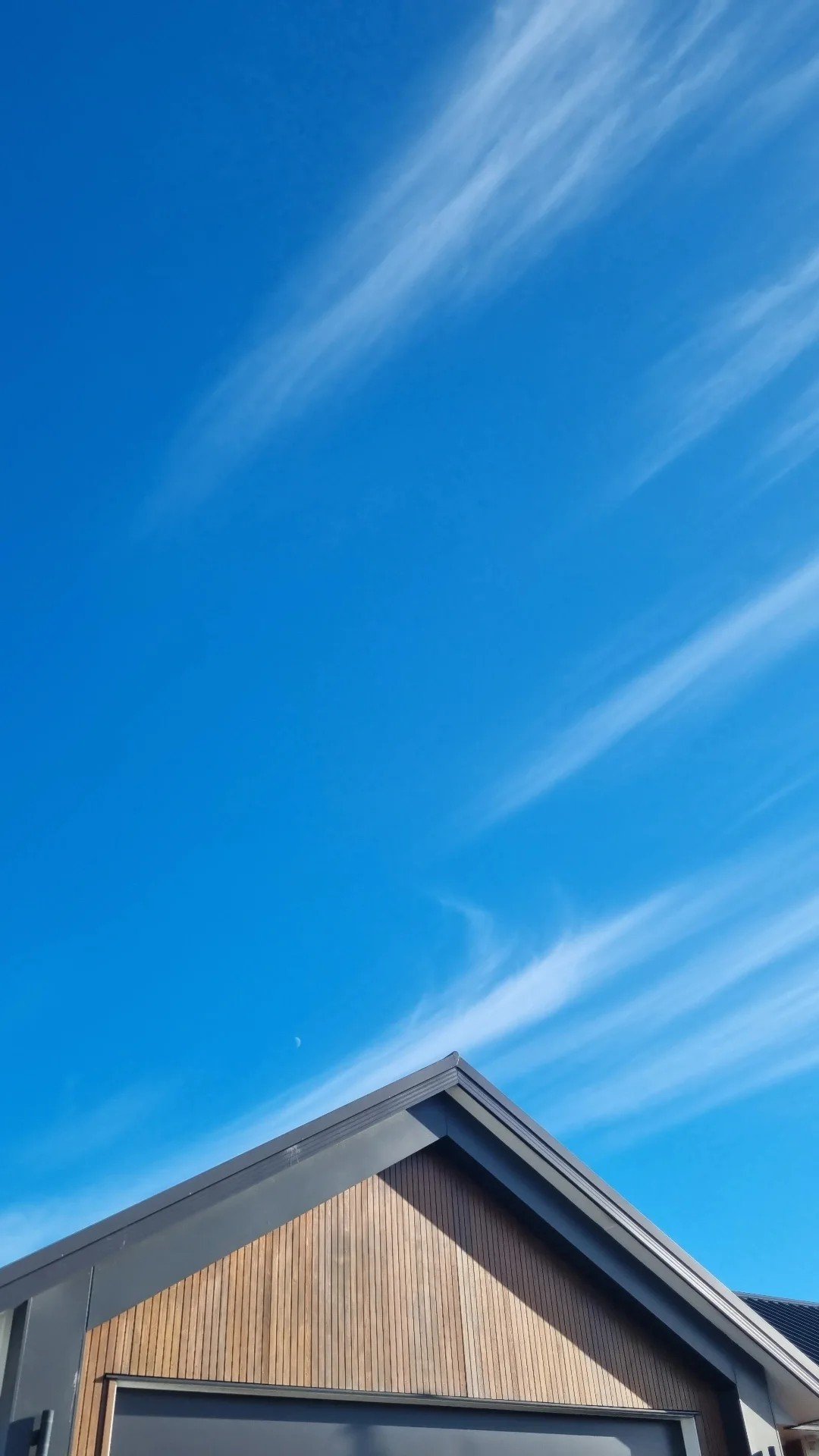Part of a house with a dark wooden exterior and gray roof against a bright blue sky with wispy clouds.