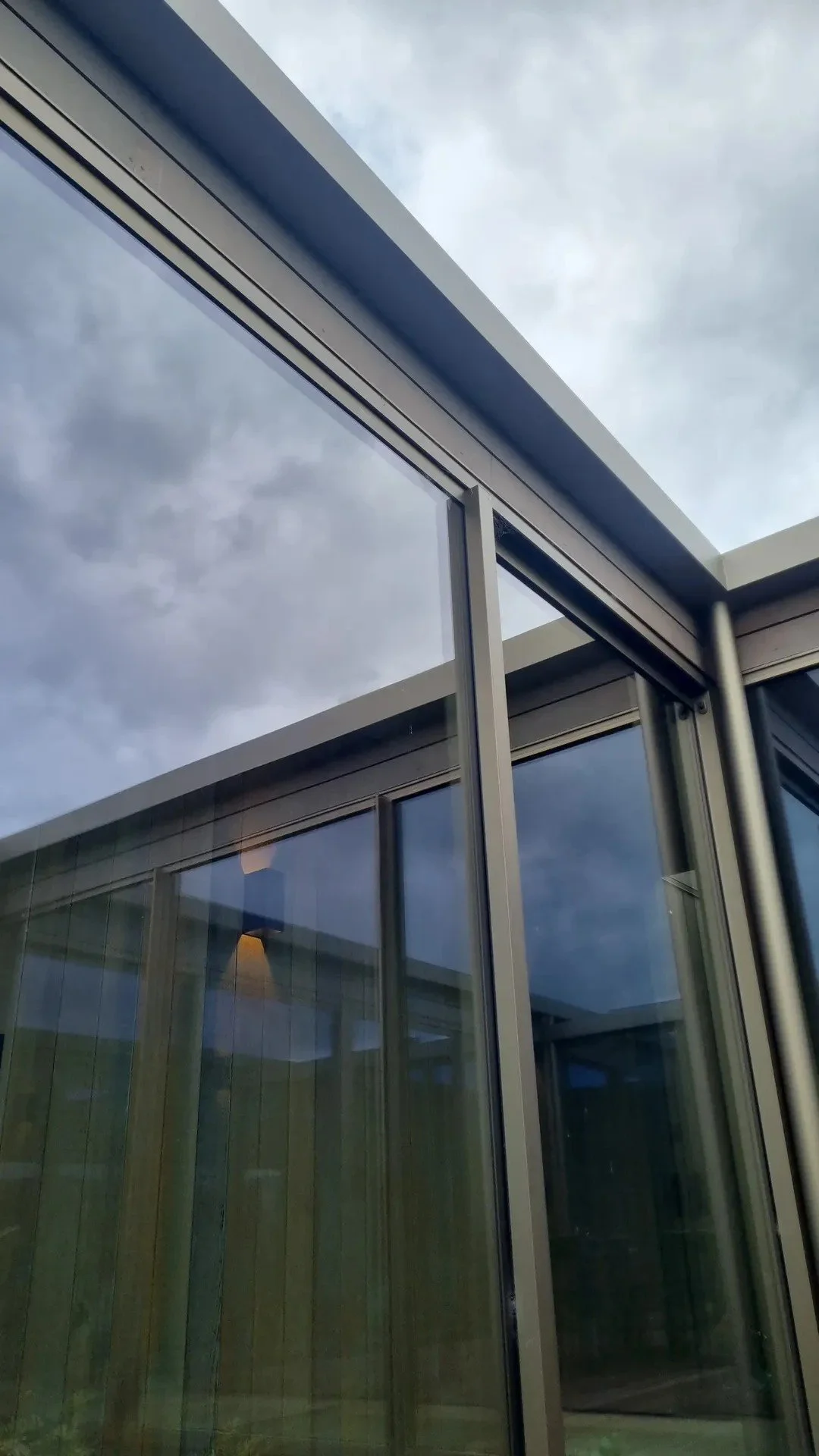 A modern building with large glass windows and metal framing, with a cloudy sky reflected in the glass.